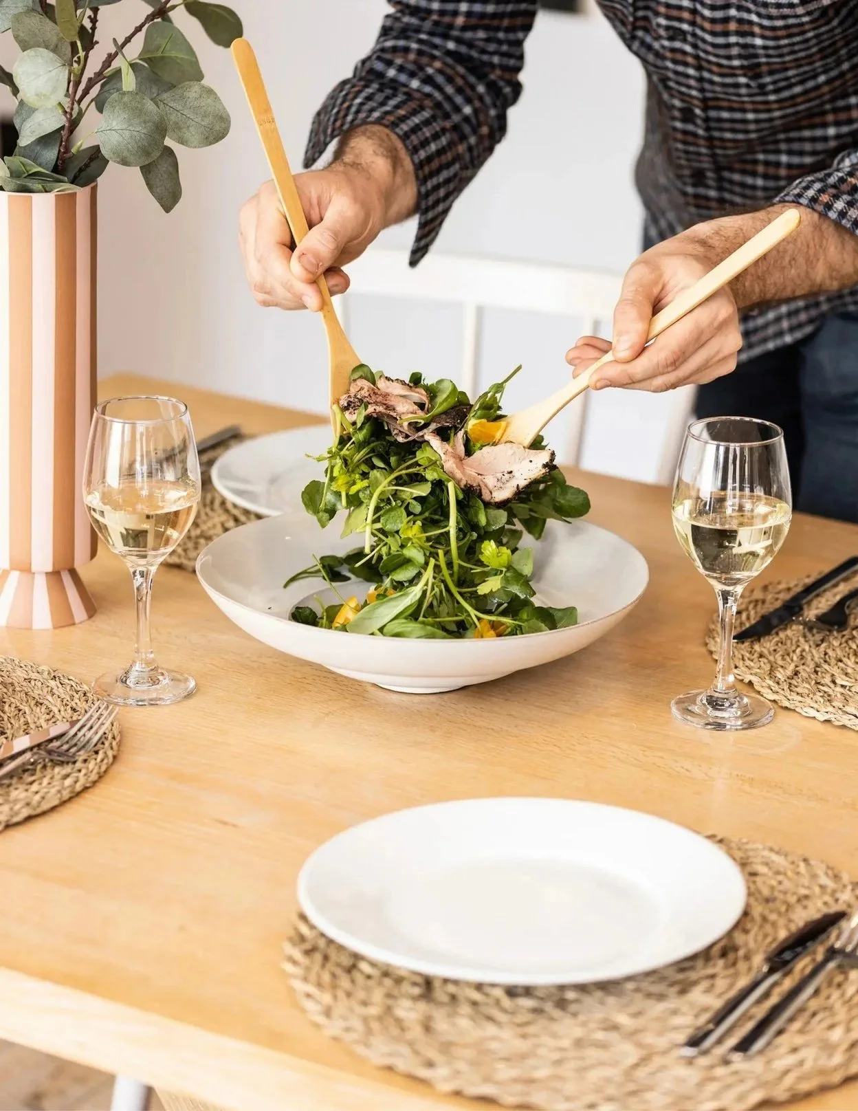 Person serving salad onto a white plate at a dinner table with two glasses of white wine, woven placemats, and a potted plant in the background.