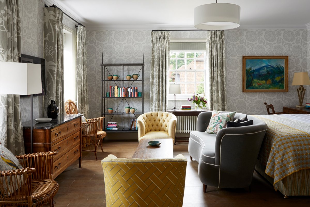 Living room with a yellow armchair, gray armchair, wooden coffee table, decorative pillows, bookshelf, dresser, with windows and curtains, and a bed partially visible in the background.