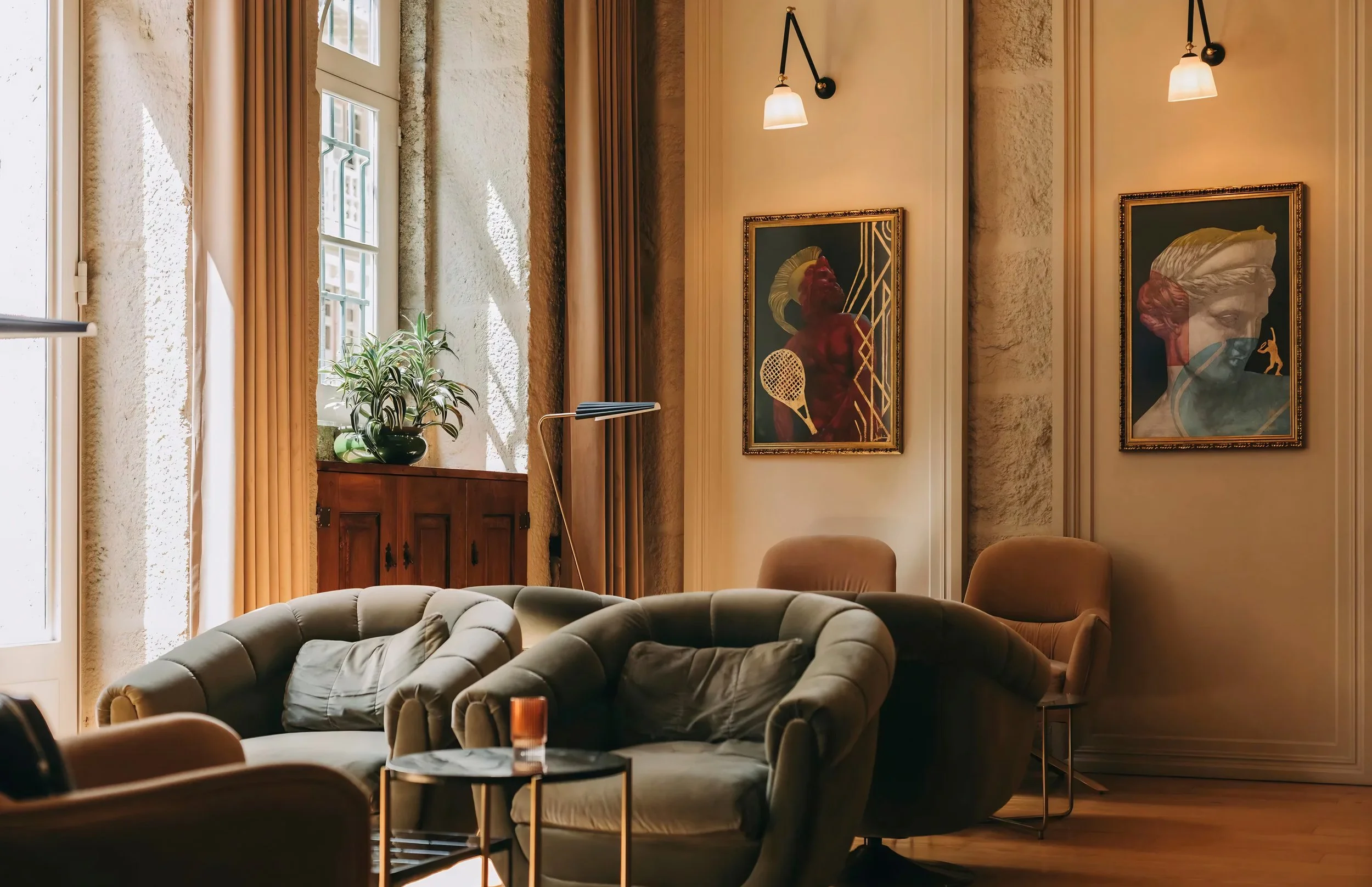 A cozy living room with large windows, beige curtains, a potted plant on a wooden cabinet, two framed art pieces on the wall, a beige tufted sofa, and a round marble coffee table with a small orange object on top.