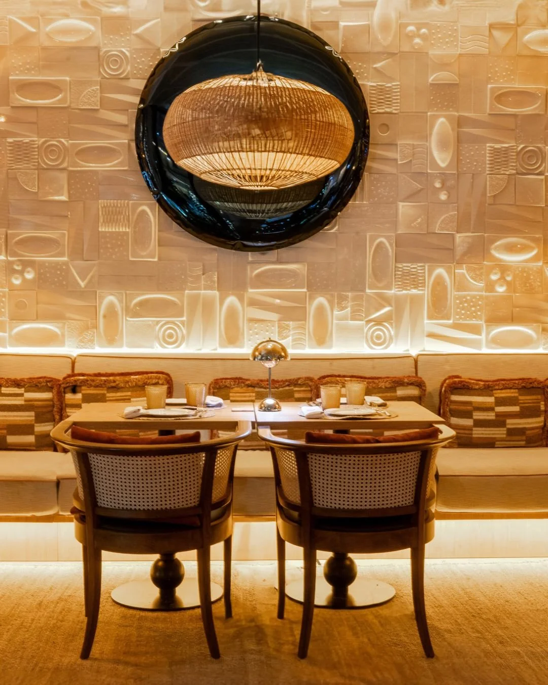 A dinner table set for two in a modern restaurant with a decorative wall, large circular mirror, and ambient lighting.