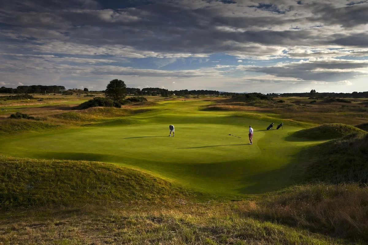 A golf course landscape with two people playing golf on the green, rolling terrain, and a wide sky with scattered clouds.