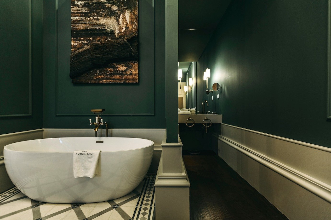 Empty bathroom with a white bathtub, green walls, and a dark wood floor. There are wall-mounted lights and a sink with a mirror in the background.