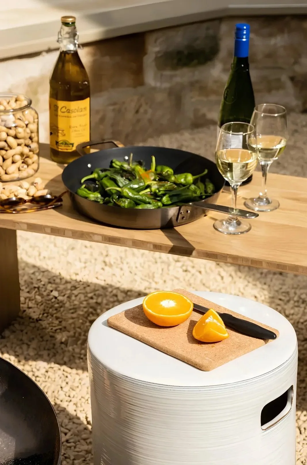 A dining scene with a wooden table holding a pan of green shishito peppers, two glasses of white wine, a bottle of white wine, a jar of mixed nuts, and a bottle of olive oil. In the foreground, a white stool has a cutting board with half an orange, a wedge of orange, and a black-handled knife.