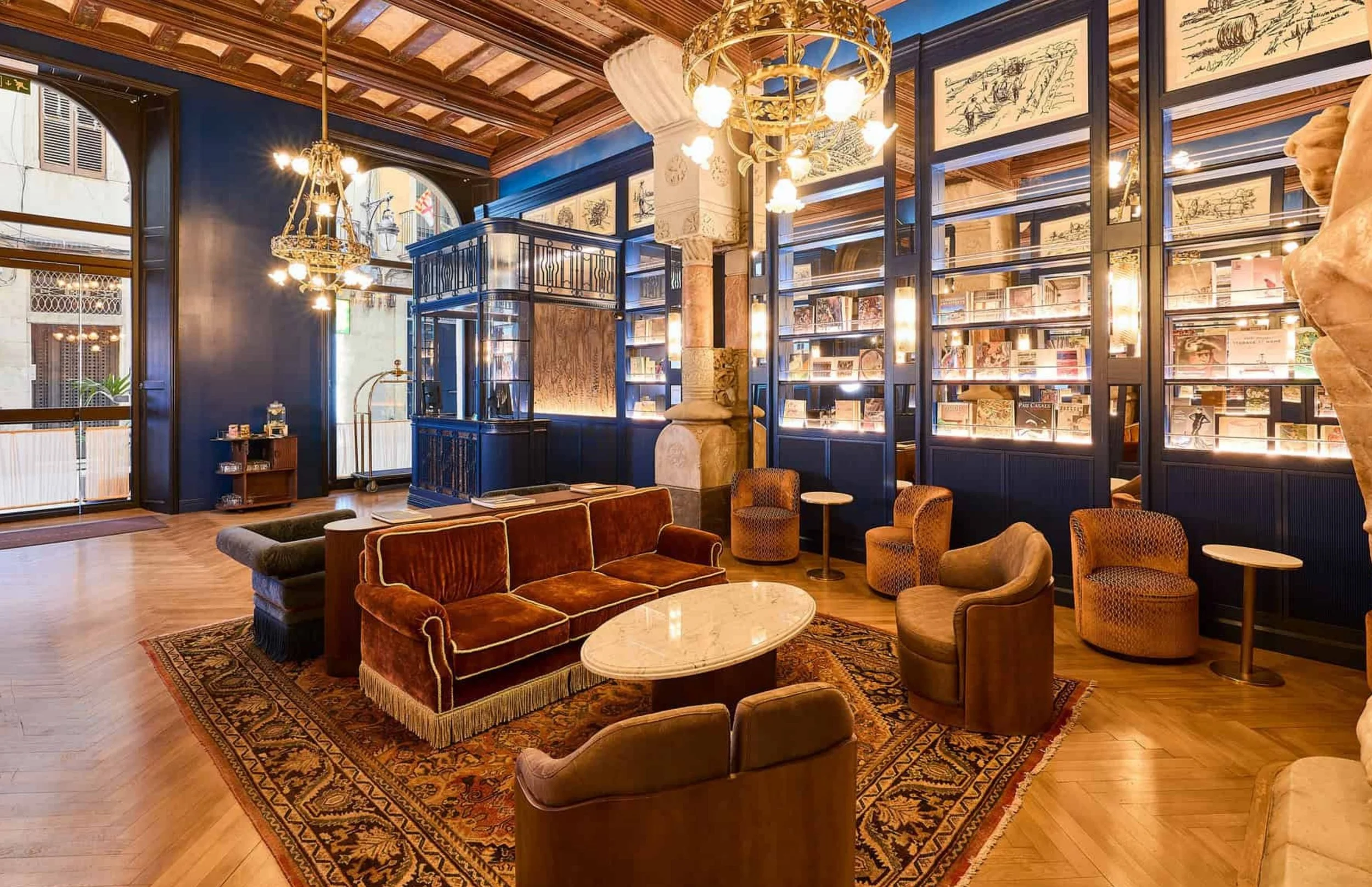 A cozy hotel lobby with warm lighting, vintage velvet and fabric chairs, a patterned area rug, wooden flooring, and large windows allowing natural light. Decorative chandeliers hang from a wood-beamed ceiling, and blue bookshelves with glass fronts d