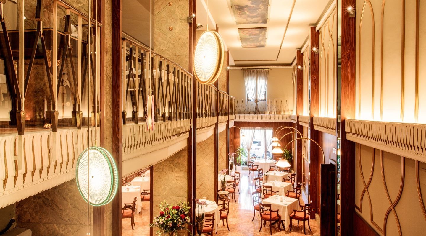 Elegant restaurant interior with tables and chairs, decorative lighting, and a balcony overlooking the lower level, featuring warm lighting and ornate wall paneling.