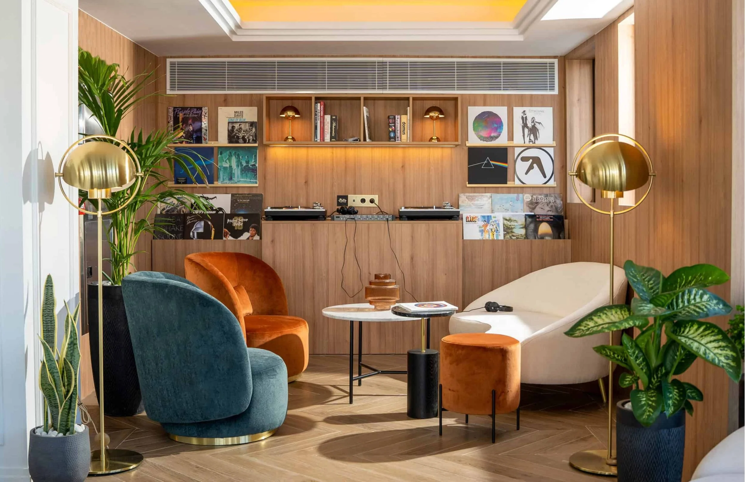 Living room with colorful velvet chairs, a white sofa, potted plants, a round coffee table with vinyl records, and wall-mounted shelves with books, vinyl, and artwork.