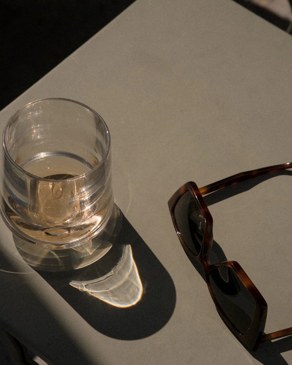 A glass of water with sunlight creating a shadow and reflection, placed on a white surface with a pair of sunglasses nearby.
