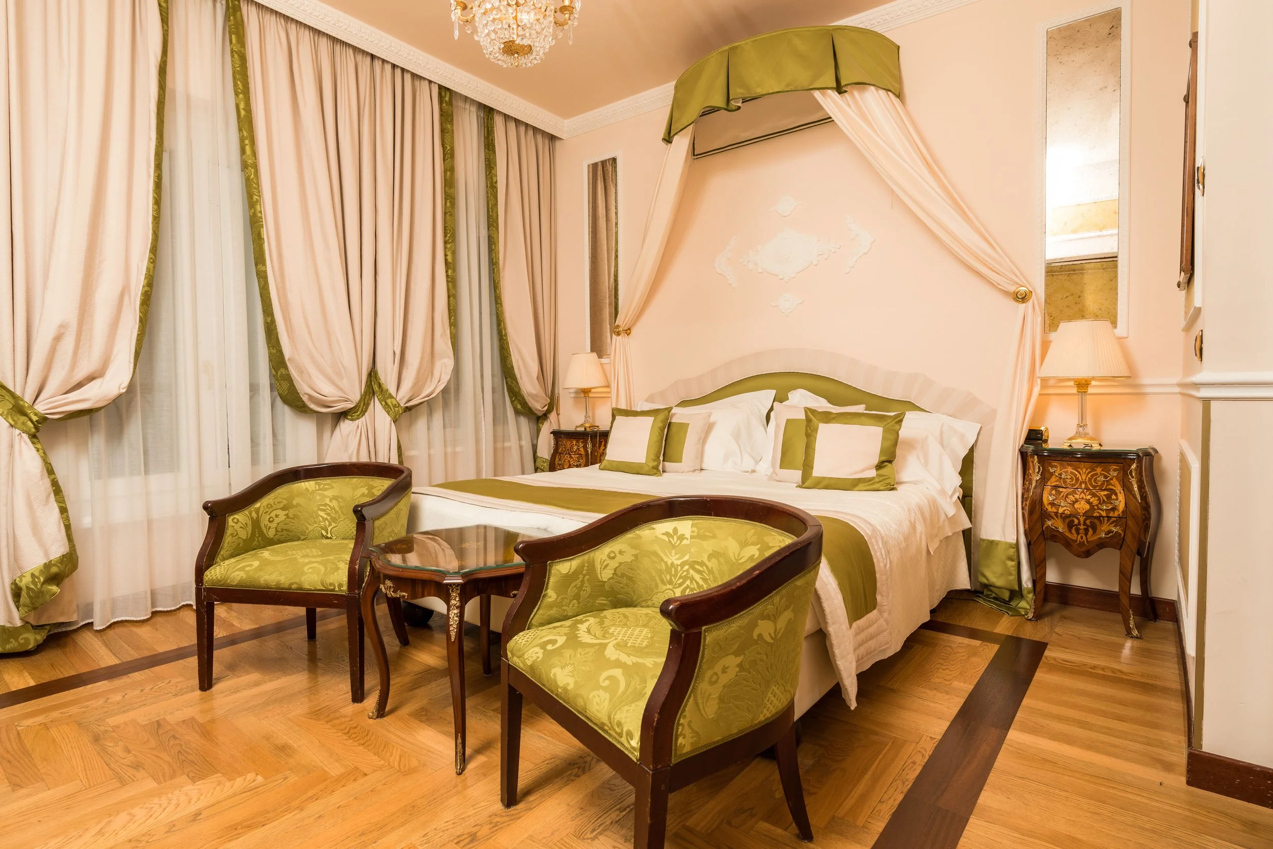 Elegant hotel room with a king-sized bed, green and cream drapes, matching pillows, vintage chairs, hardwood floors, and chandeliers.