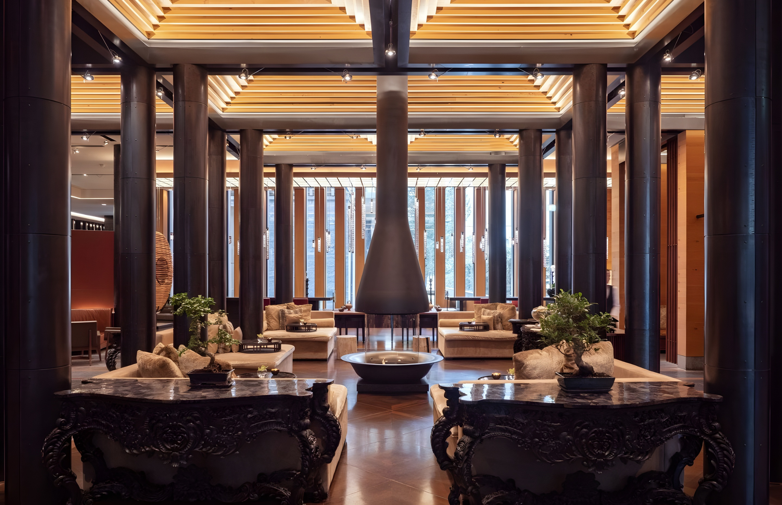 Interior of a modern hotel lobby featuring tall cylindrical dark wood pillars, a central fireplace, plush chairs, potted plants, and large windows with vertical wooden decorative elements.