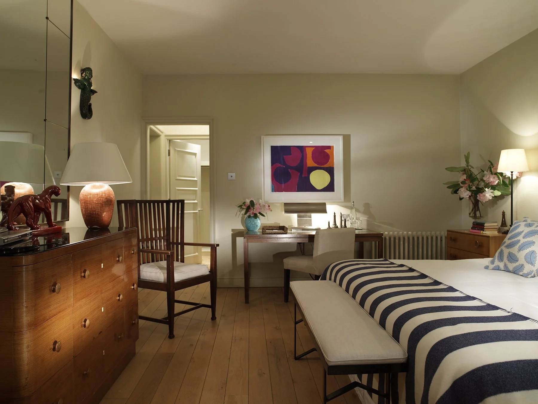 A cozy bedroom with a wooden dresser, a nightstand with pink flowers, a framed abstract art piece, a desk with a beige chair, a lamp, and a bed with a striped blanket and cloud-patterned pillow.
