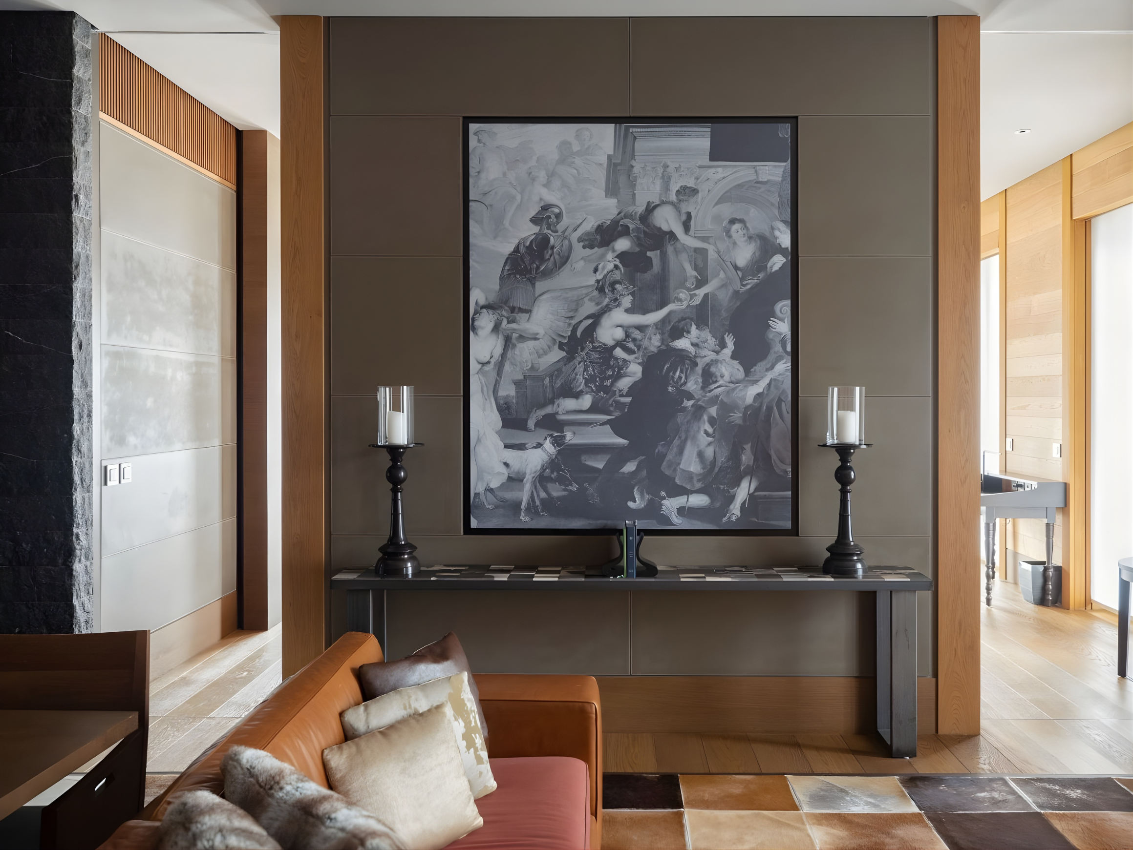 Interior shot of a modern living space featuring a large black and white classical painting with sculptures and figures in the background, flanked by two black candlesticks with white candles, a brown leather sofa with pillows, and wall paneling with wood accents and large windows in the background.