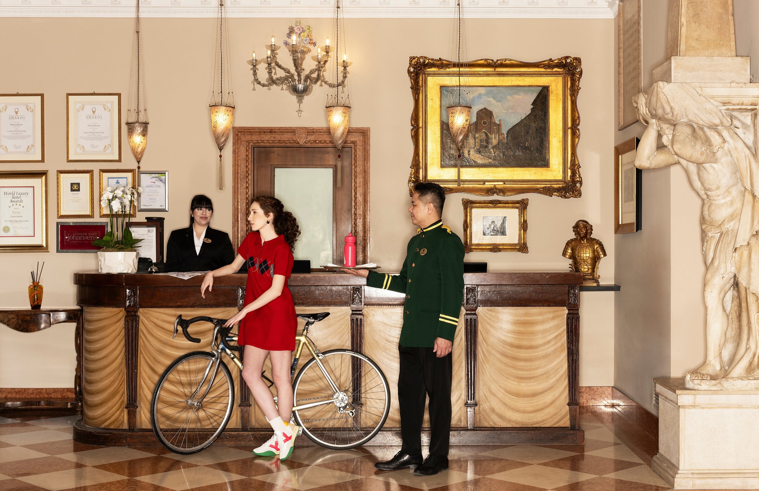 Hotel reception area with a young woman in red dress with cycling shoes and a bicycle, standing beside a hotel staff member in uniform. The staff member is holding a red thermos and a tray, while another staff member looks on. The background features