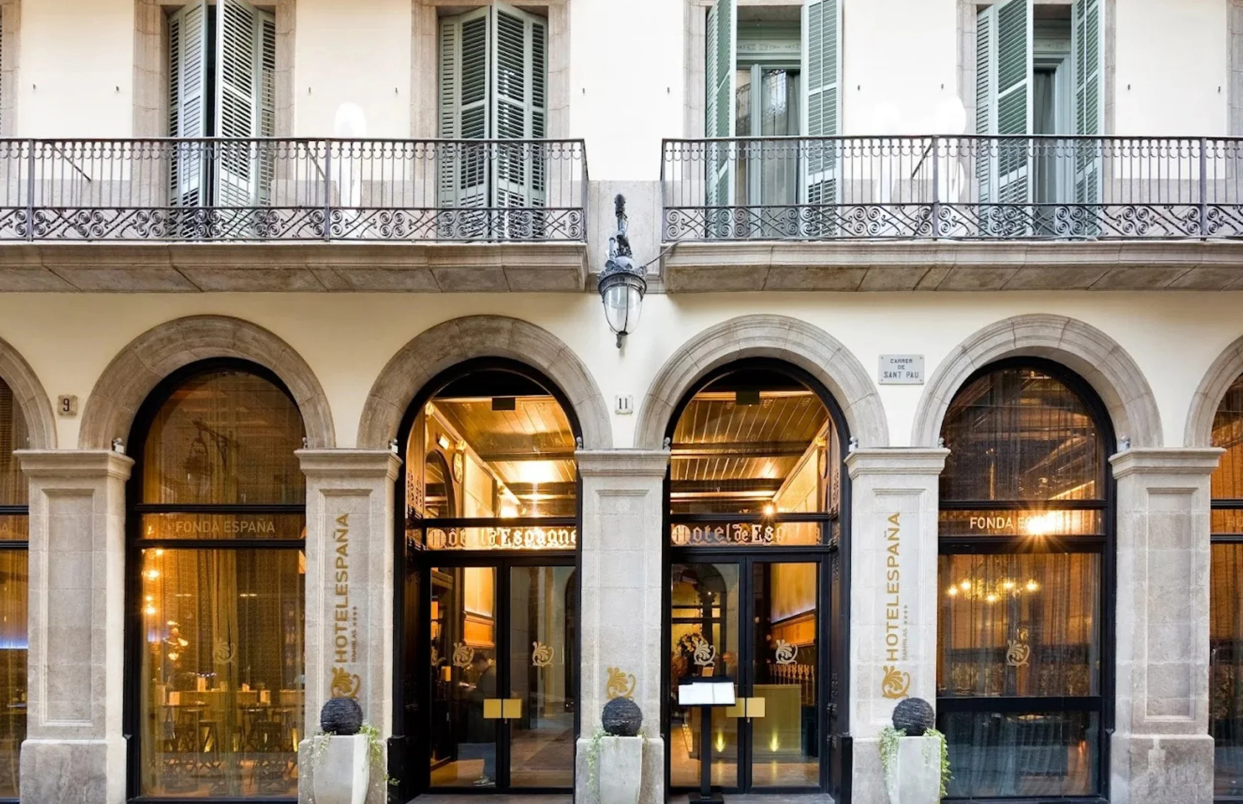 Facade of a hotel with large arched windows and a balcony with ornate iron railings, a street lamp, and a sign indicating street name 'Carrer de Sant Pau'.