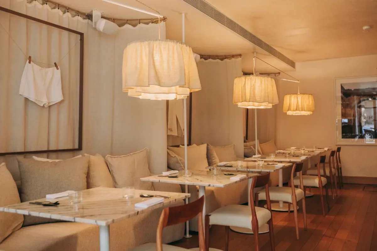 Empty restaurant with beige couches, tables, wooden chairs, hanging lamps, and a wall art with white clothing.