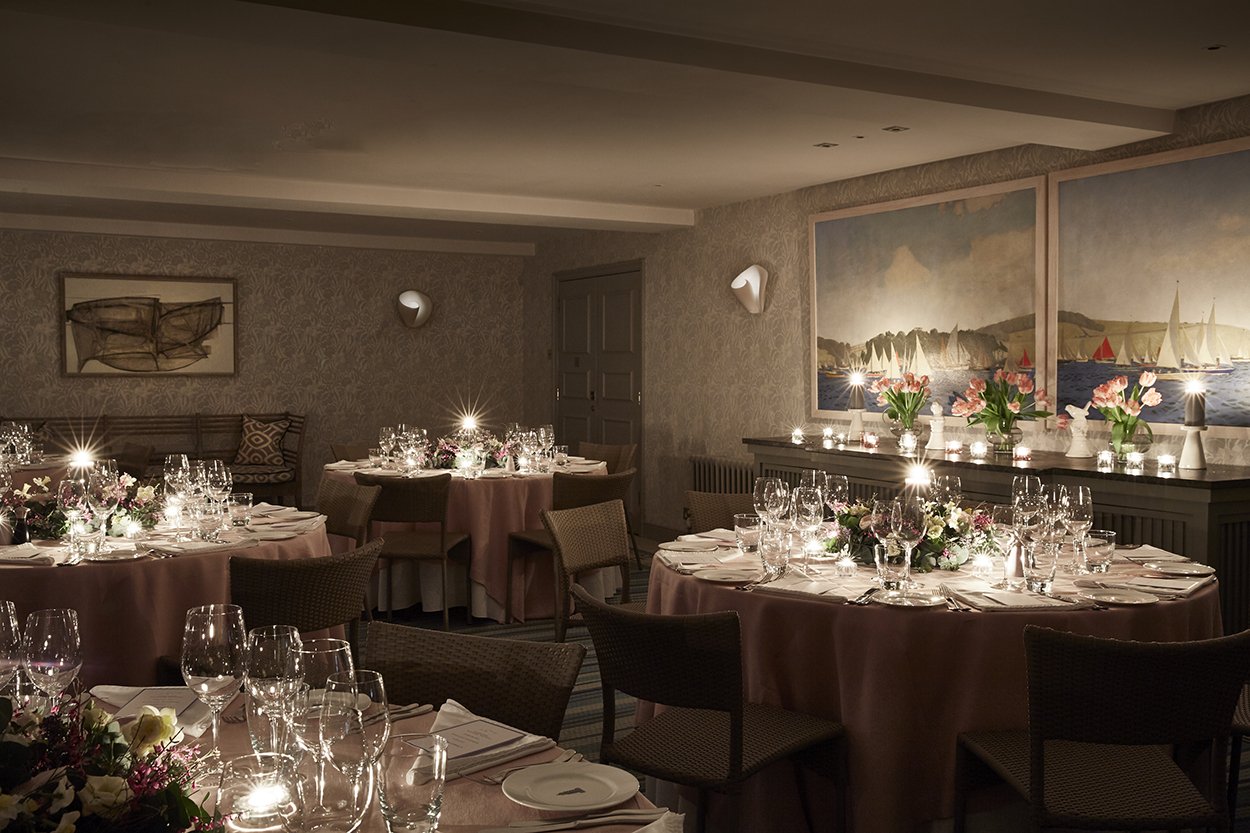 A dimly lit banquet room with round tables covered in pink tablecloths, decorated with floral centerpieces and surrounded by chairs, set for a formal dinner with numerous wine glasses, cutlery, and plates. The walls have paintings and subdued lighting.