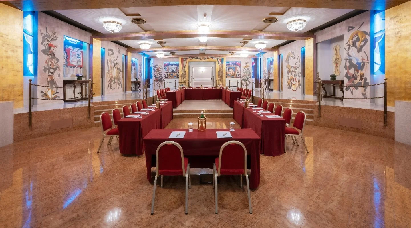 A large conference or banquet room with red tables and chairs arranged in a U-shape. The room features ornate murals on the walls, chandeliers on the ceiling, and a raised platform at the far end.