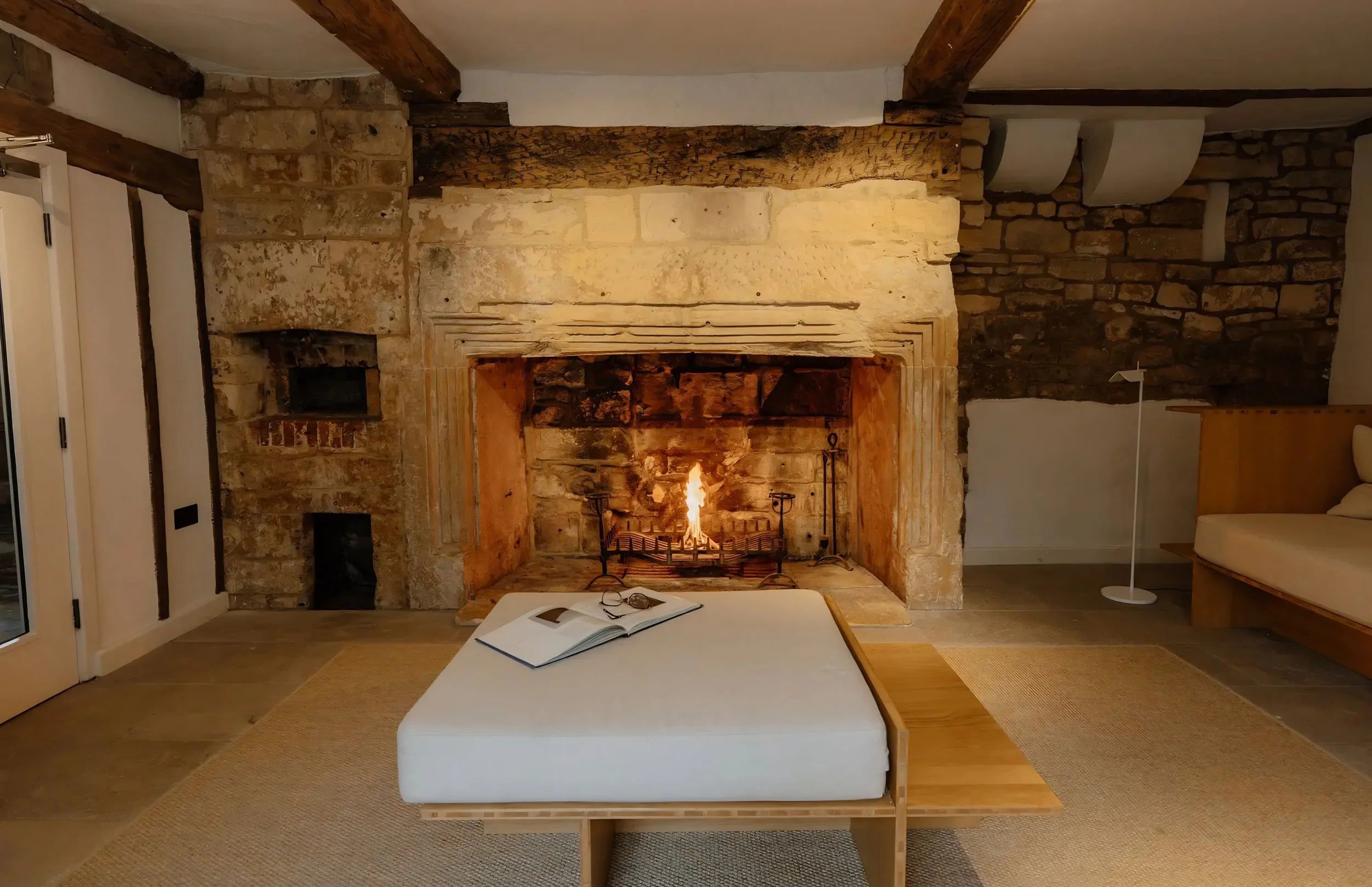 A cozy living room with a stone fireplace lit with a fire, a white ottoman with an open book and glasses on it, and a wooden bench on a beige rug. There are wooden beams on the ceiling and brick and stone wall accents.