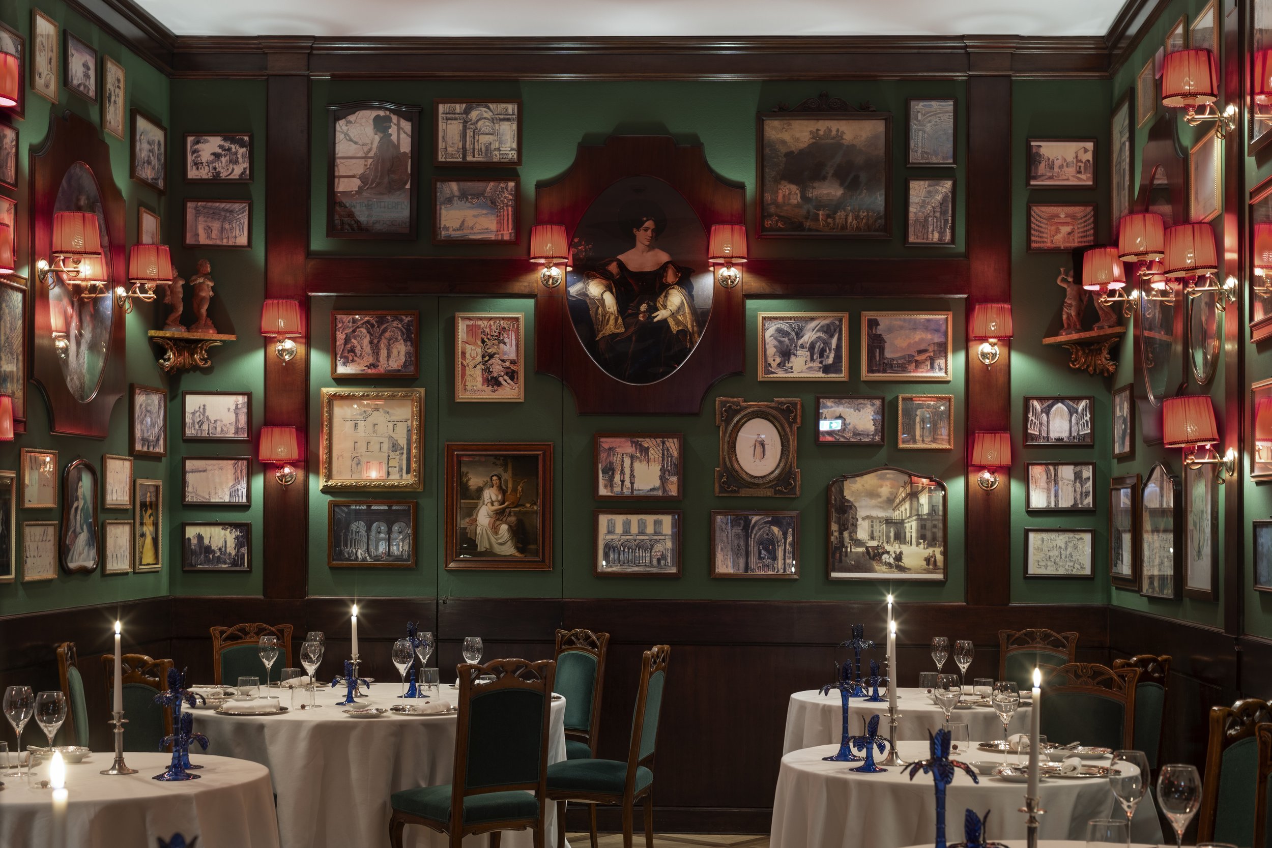 Elegant restaurant dining room with green walls, dark wood wainscoting, and a gallery wall of framed artwork and paintings. Round tables are set with white tablecloths, wine glasses, and tall candlesticks with blue decorative accents. Wall sconces wi