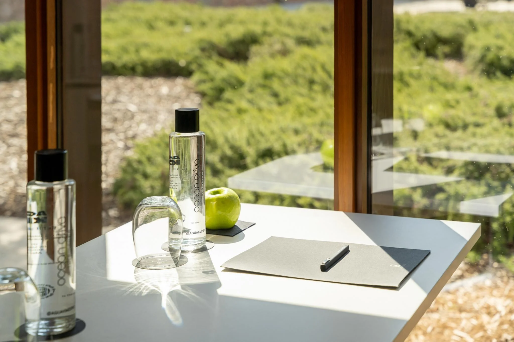 A white table with bottles of skincare products, a green apple, a glass, a notebook, and a pen inside near a window overlooking a green landscape.