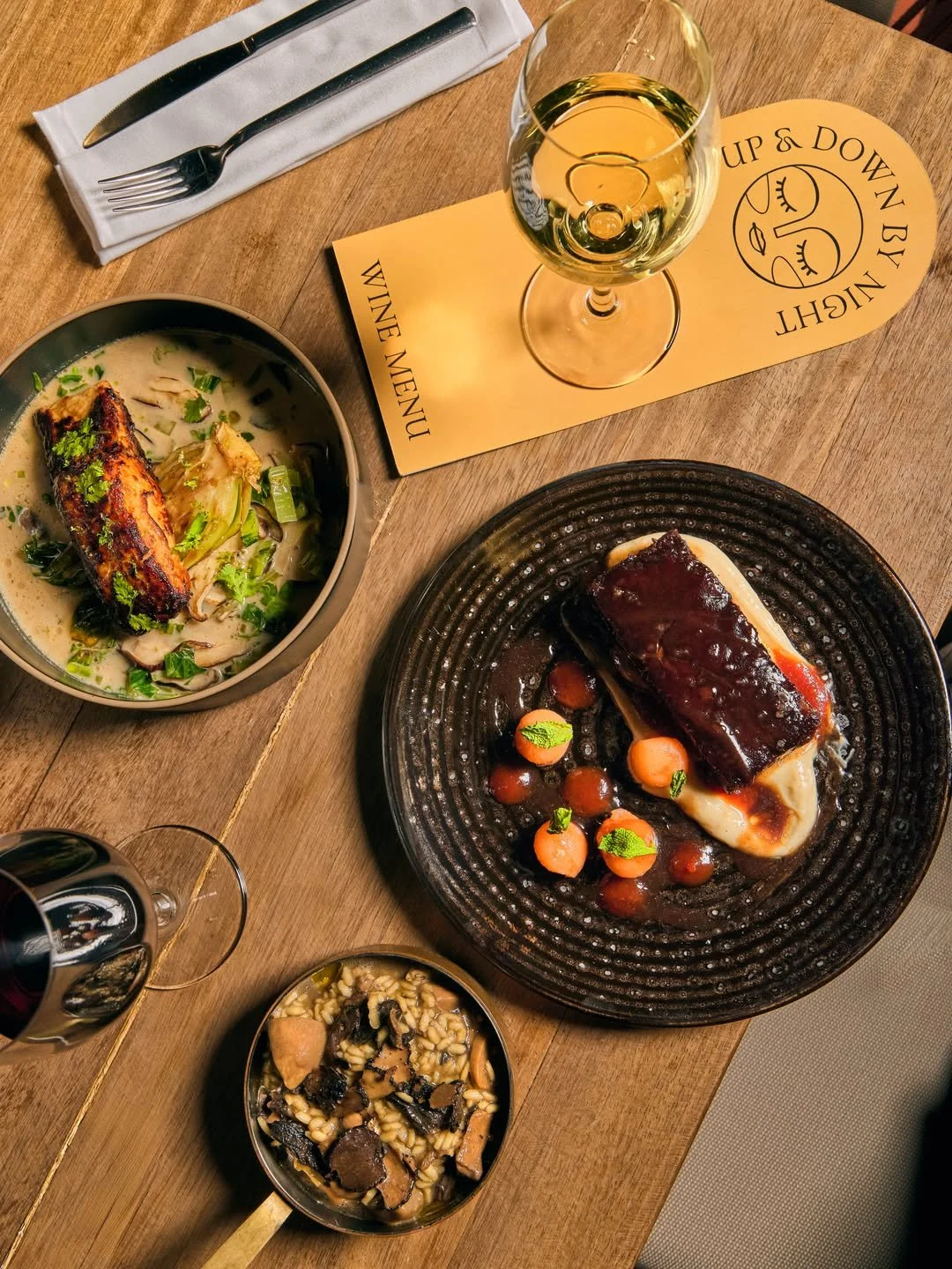 A heart-shaped steak with sauce, baby carrots, and green herbs on a black plate, a bowl of rice with mushrooms and black truffles, a bowl of creamy soup with fish, and a glass of white wine on a wooden table with a paper placemat and silverware.