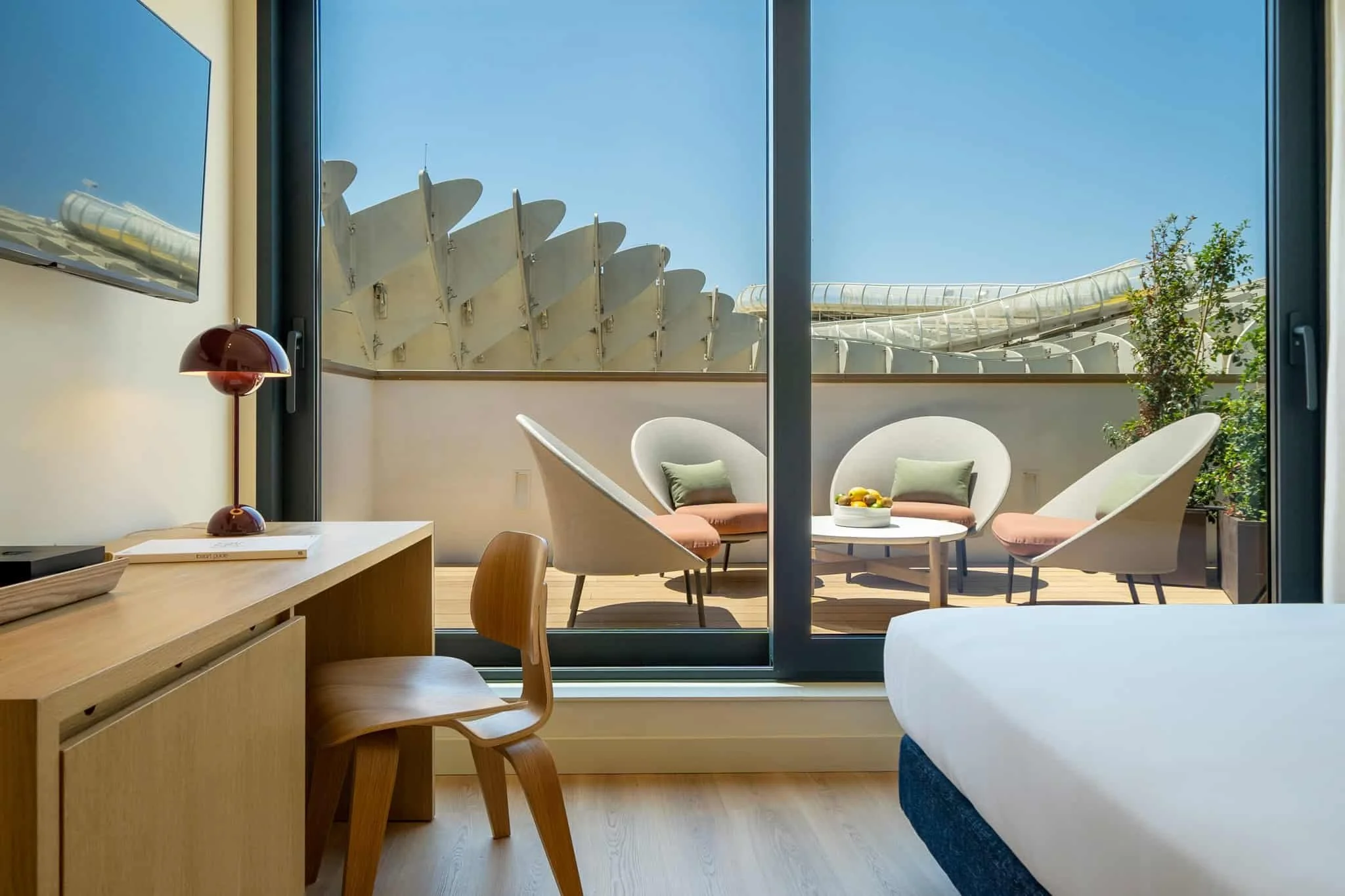 View from inside a room incorporating a sliding glass door revealing a balcony with outdoor seating, including two white chairs with cushions and a small round table with a bowl of lemons, and exterior architectural elements against a clear blue sky.