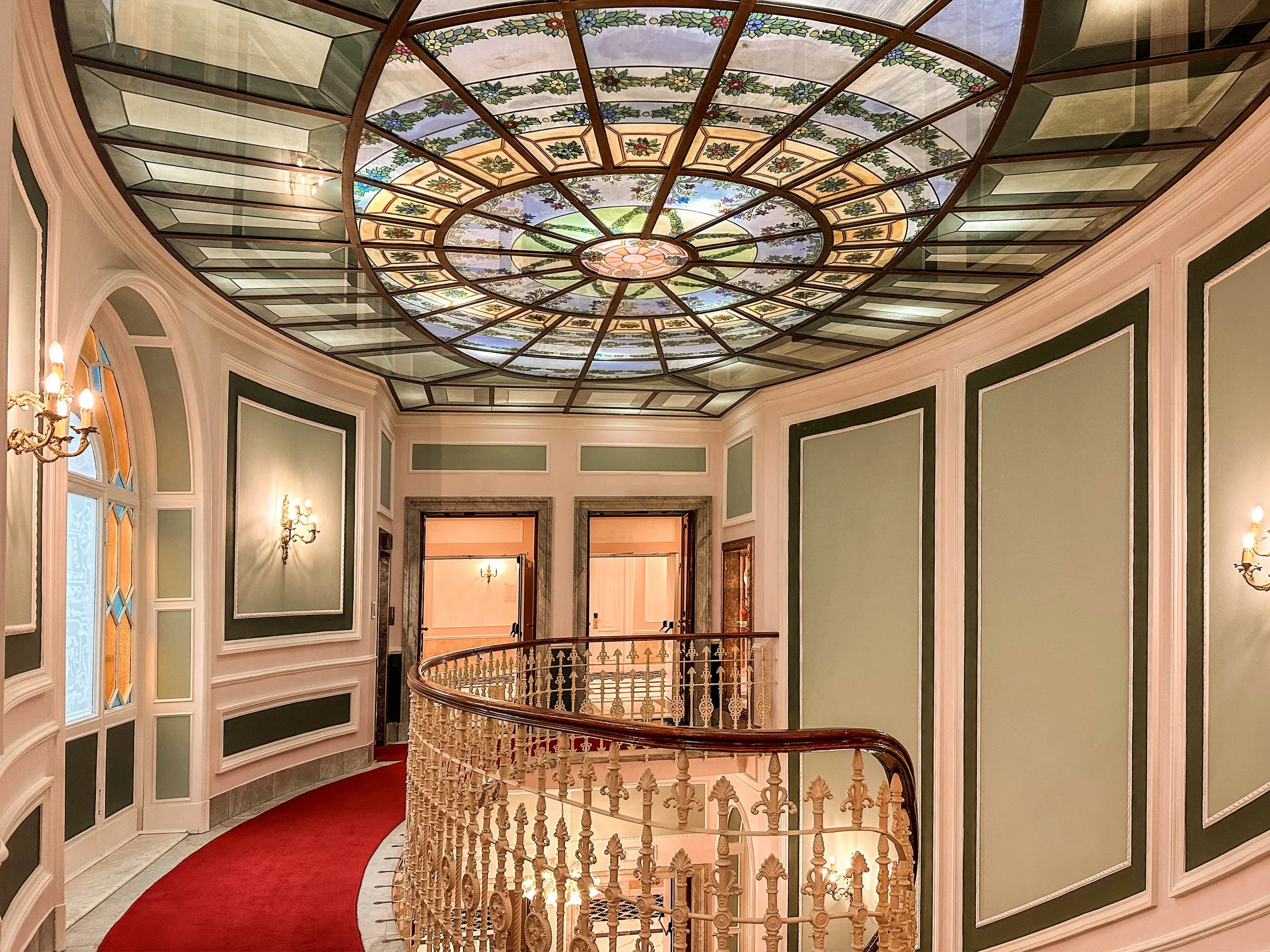 Interior of a building with ornate stained glass ceiling, framed walls with decorative molding, and elegant wall sconces, featuring a curved red carpeted staircase.