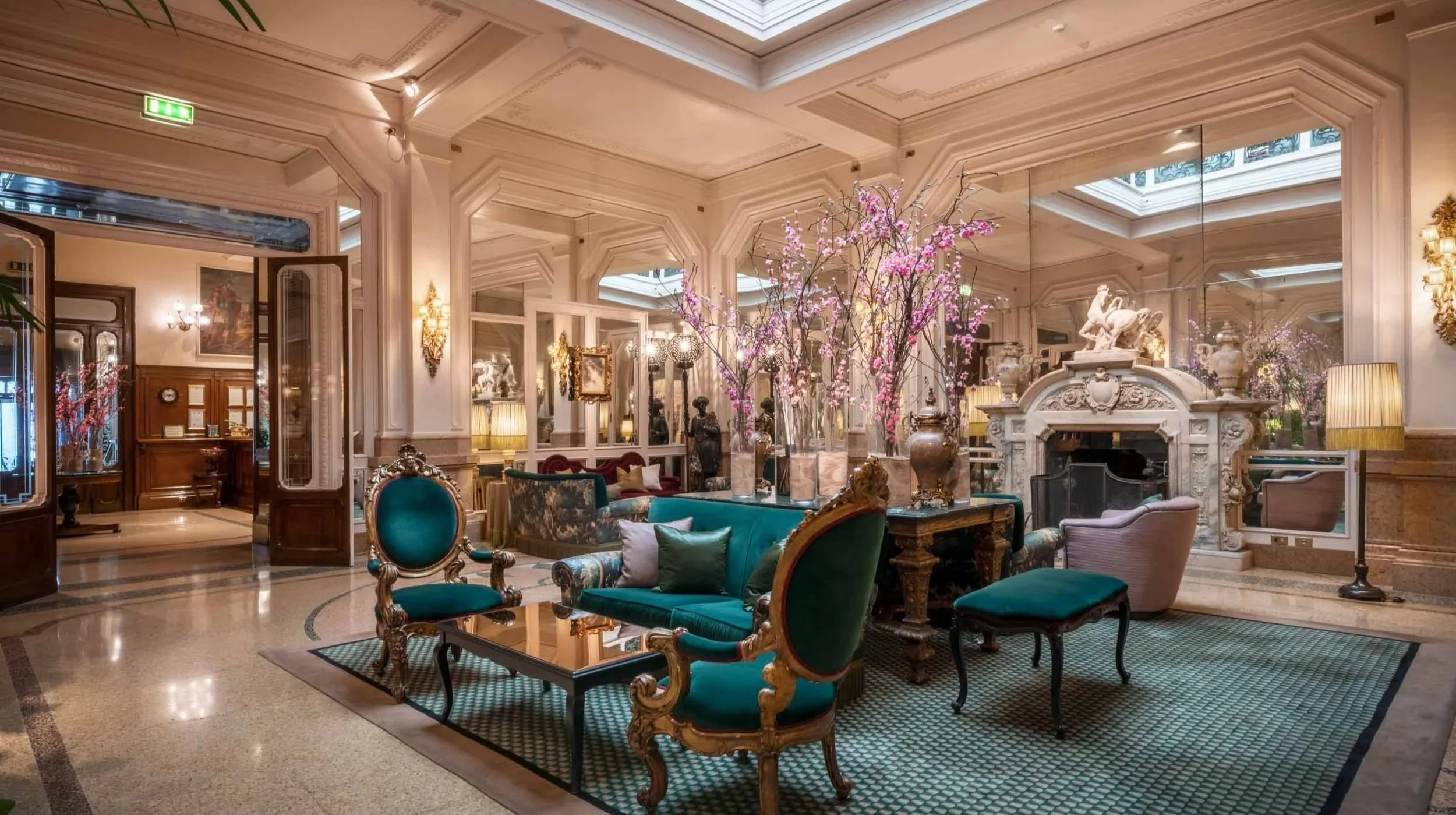 Luxurious hotel lobby with ornate ceiling, marble fireplace, pink flowering branches, elegant furniture, and decorative lighting.