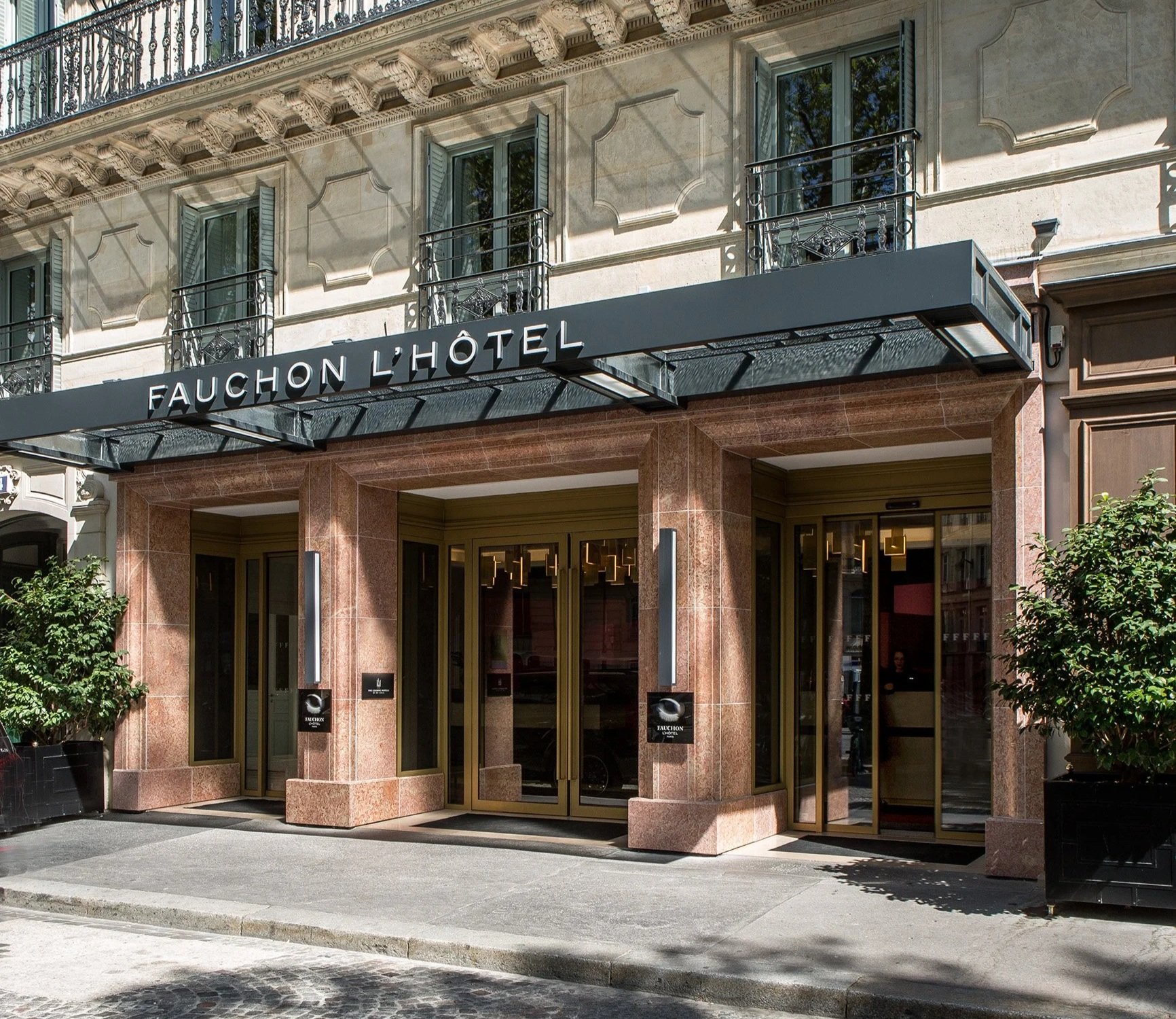 The exterior of the Fauchon L'Hôtel building features a beige stone facade, large glass double doors at the entrance, and a black canopy with white lettering. There are two small trees on either side of the entrance, and the building has several windows with decorative wrought iron balcony railings. Shadows from nearby trees cast onto the building.