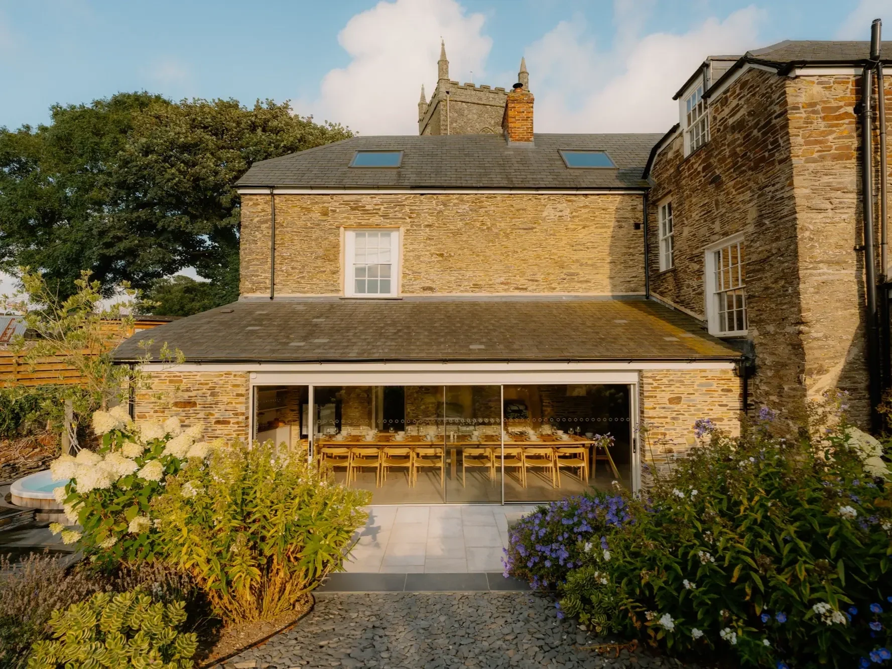 A brick house with a gray roof, large windows, and a patio with wooden chairs and a glass wall, surrounded by lush greenery and flowering plants, with a church steeple visible in the background under a partly cloudy sky.