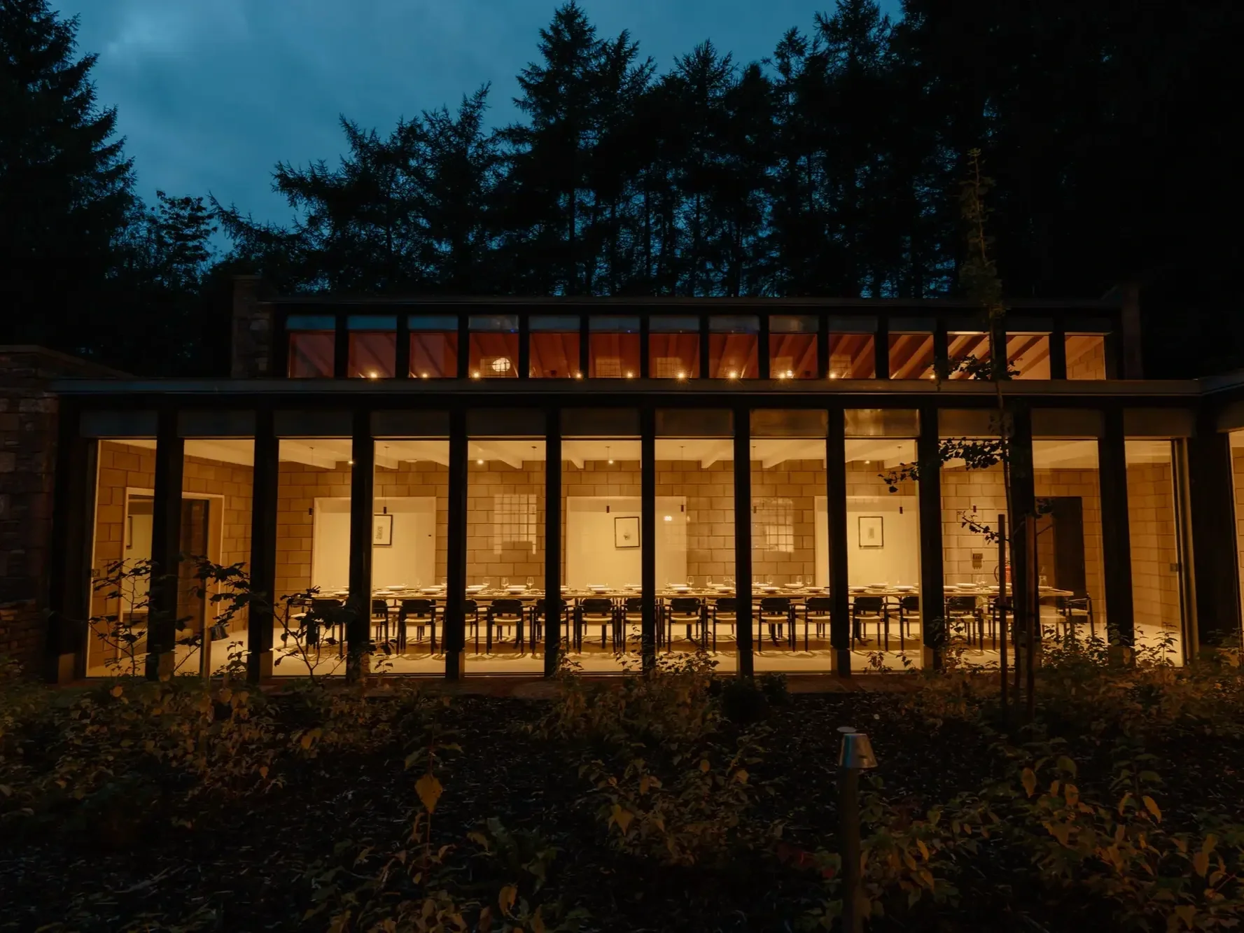 Night view of a modern building with large glass windows, warmly lit interior dining area with tables and chairs, surrounded by trees and foliage.