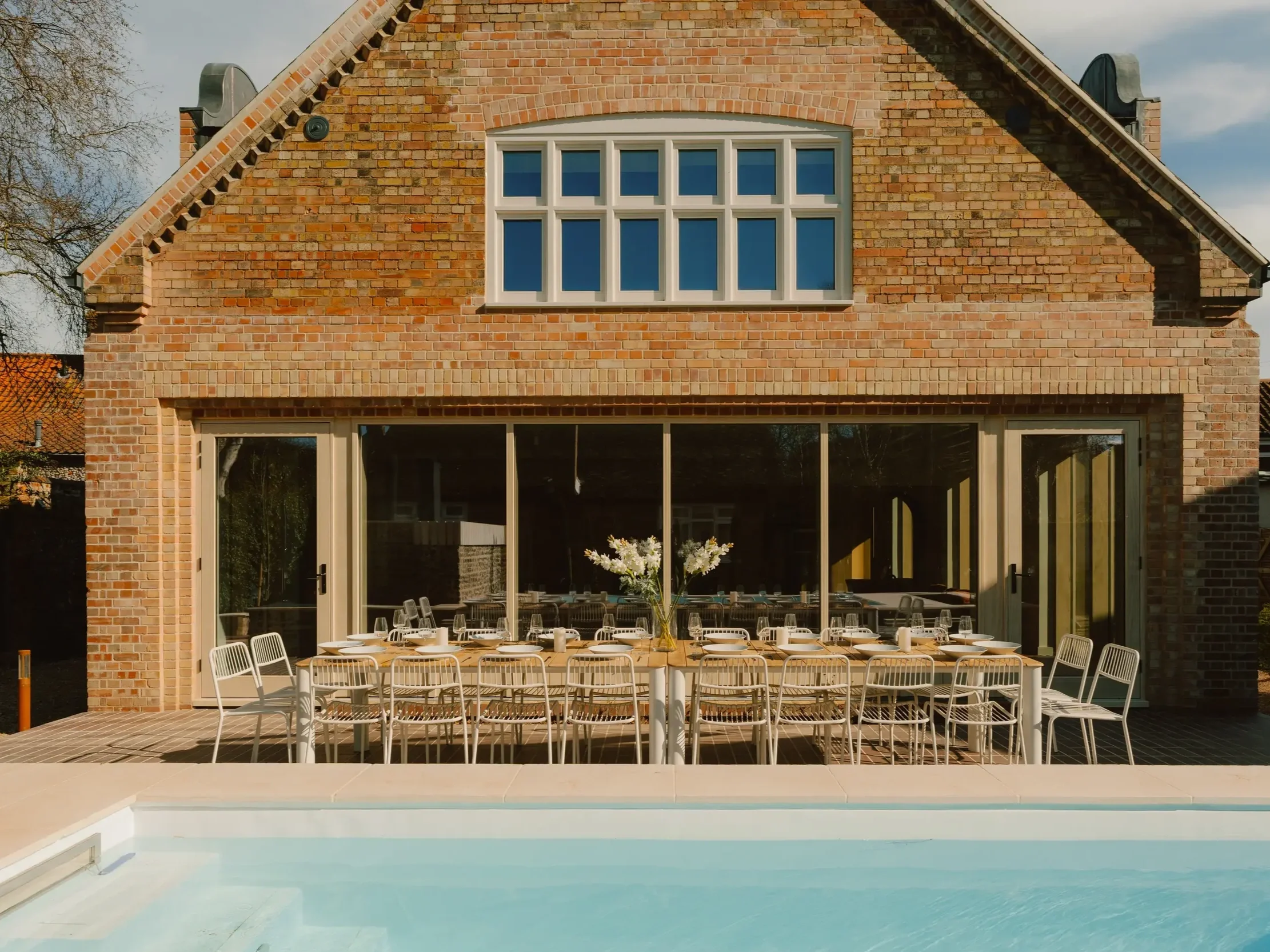 Backyard patio with a modern brick house featuring a large window. The patio has a dining arrangement with a long table, white chairs, and tableware, overlooking a swimming pool with a light blue interior.
