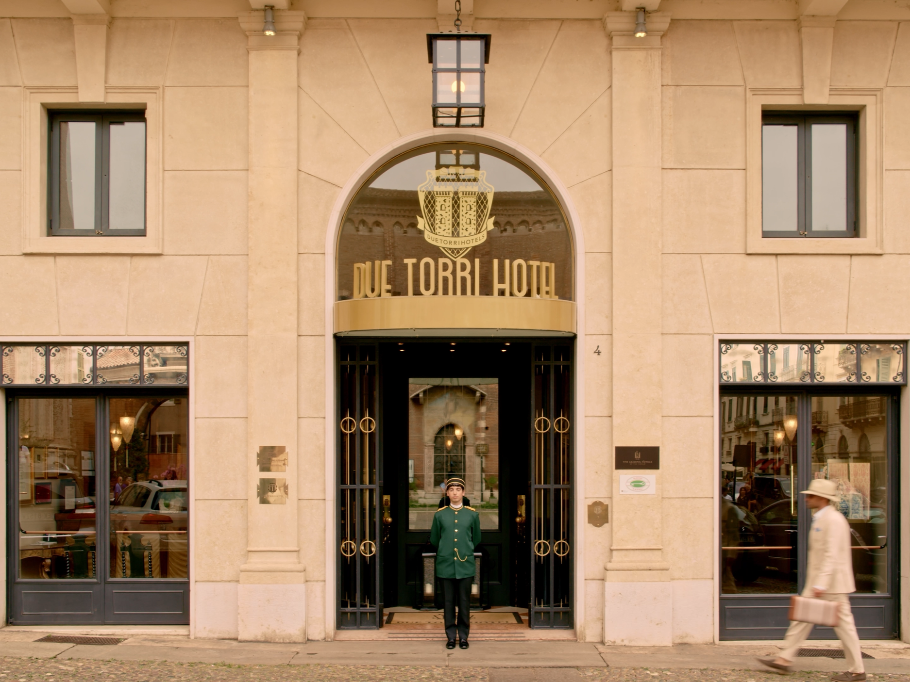 The entrance of a hotel with a large glass door and the name 'DUE TORRI HOTEL' above it. A uniformed doorman is standing at the door, and two pedestrians are walking by on either side. The building has a beige stone facade with three large windows above the entrance, and a hanging lantern light fixture.