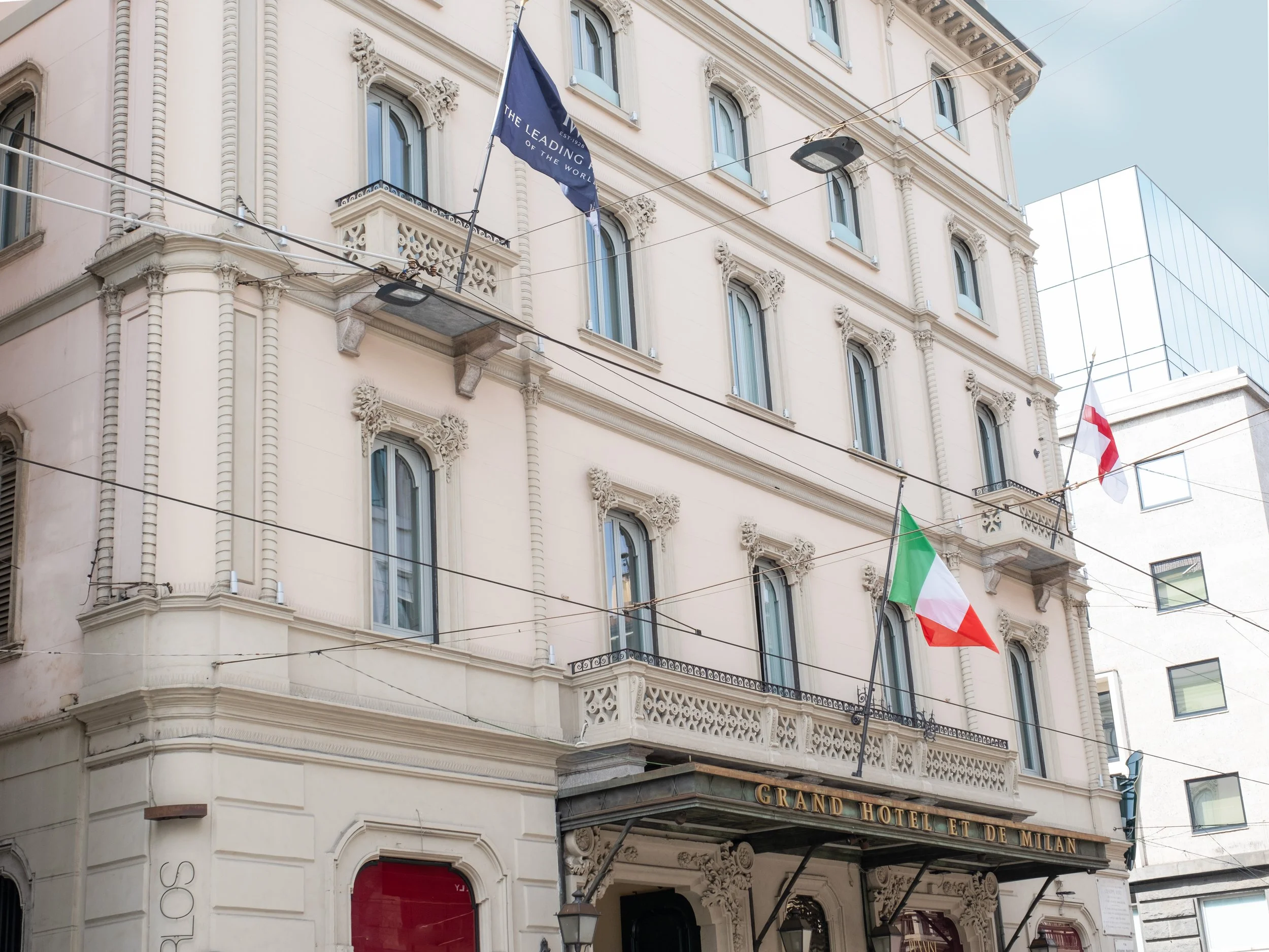 Facade of a historic building with ornate architectural details, balconies, and flags, including the Italian and Swiss flags, in an urban setting.