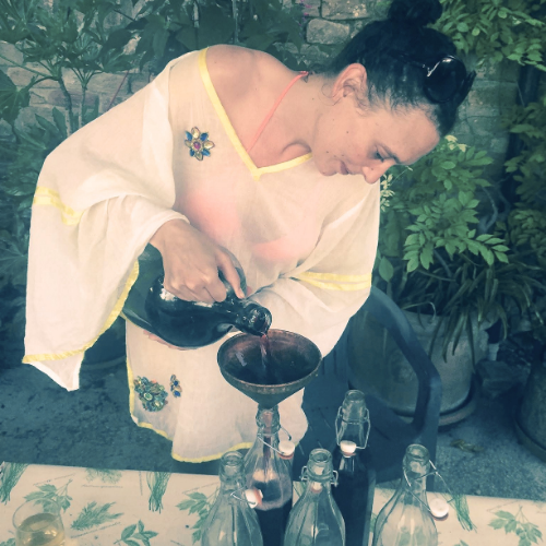 A woman in a yellow dress pouring a dark beverage from a bottle into a glass bottle on a table with other glass bottles, outdoors with green plants.