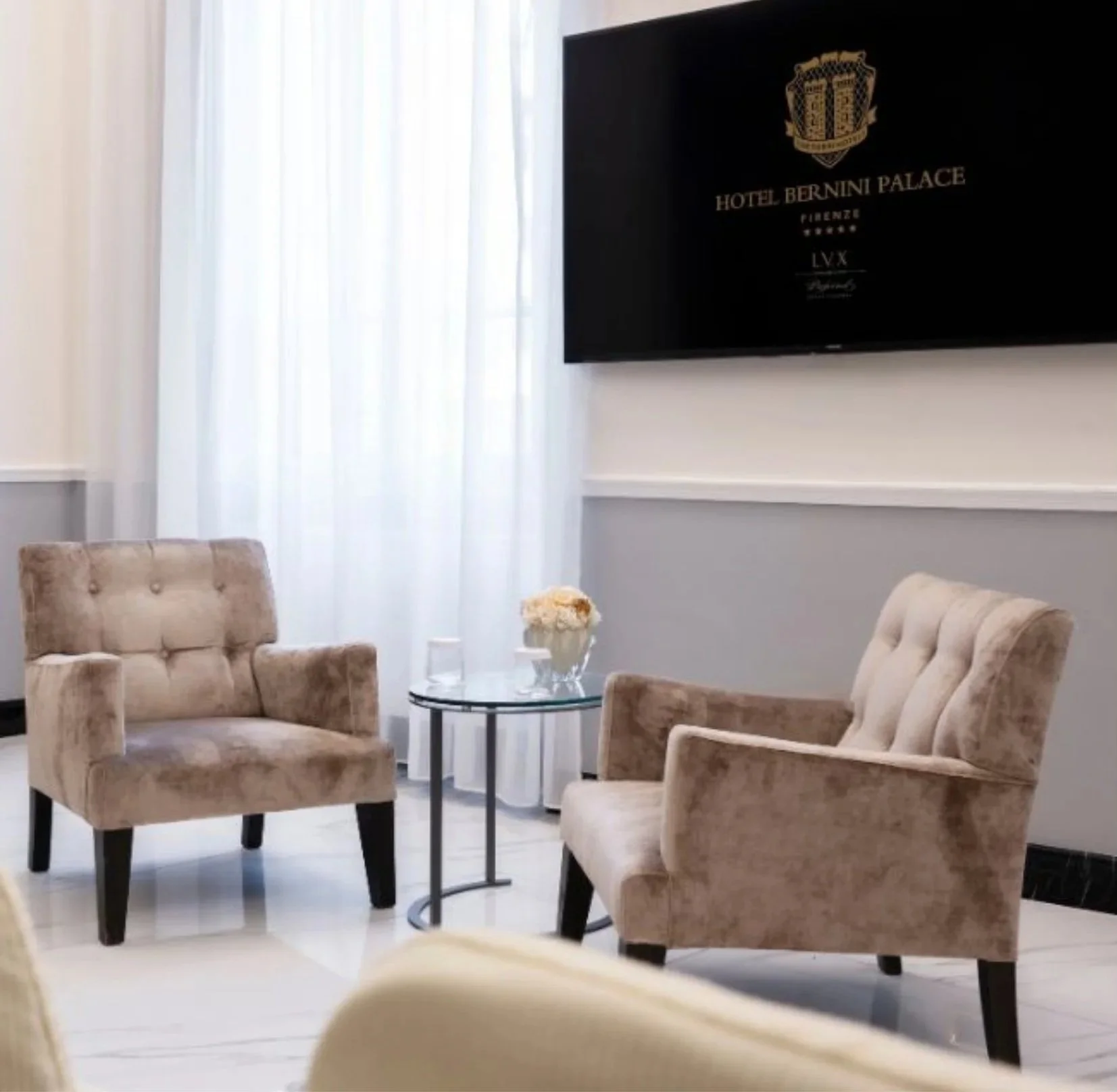 A hotel lounge area with beige velvet armchairs, a glass-top round table with a white flower arrangement, white curtains, and a wall-mounted flat-screen TV displaying the Hotel Bernini Palace logo.