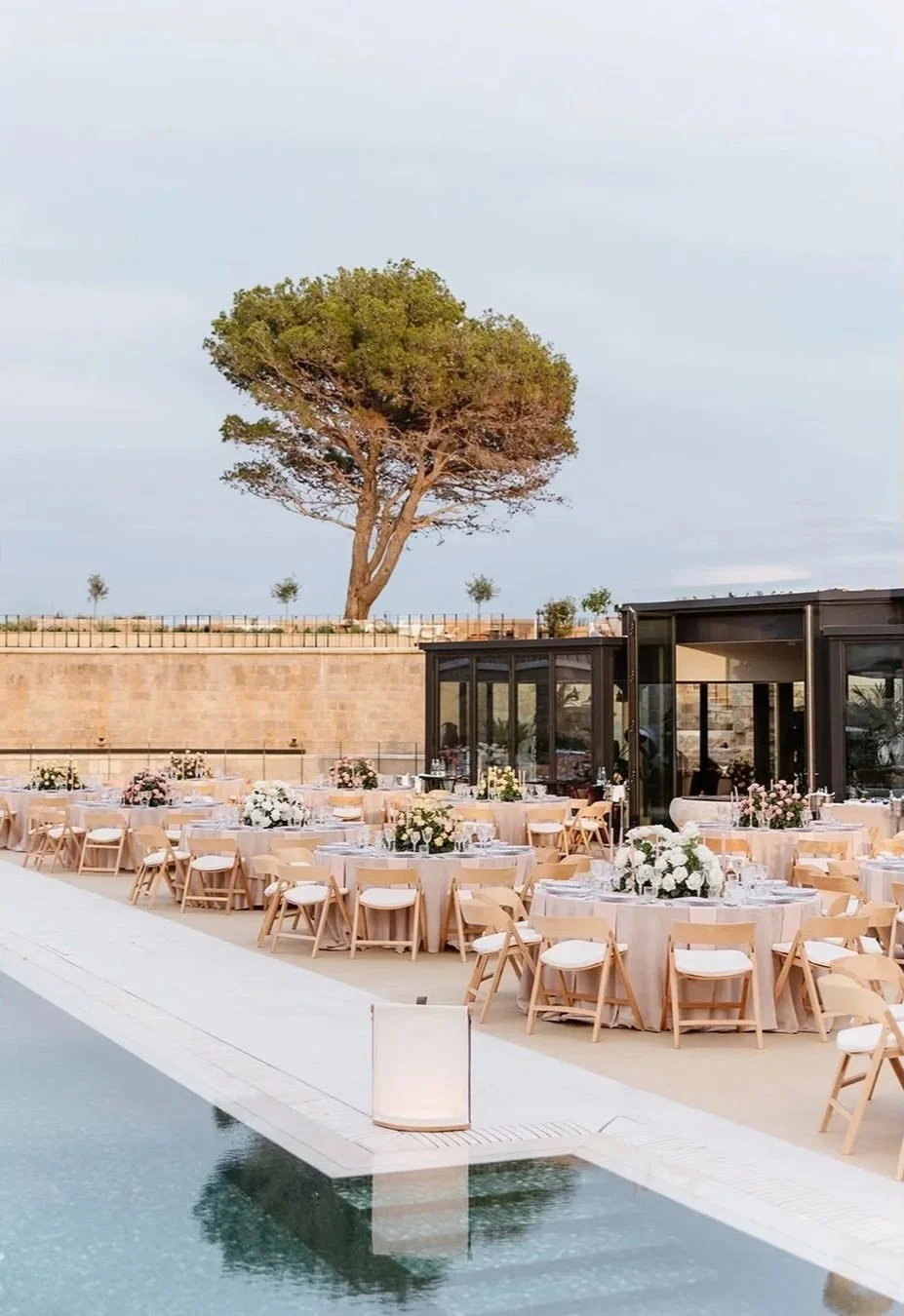 Outdoor event setup with round tables decorated with floral centerpieces, surrounded by wooden chairs, overlooking a pool with a large tree in the background under a partly cloudy sky.