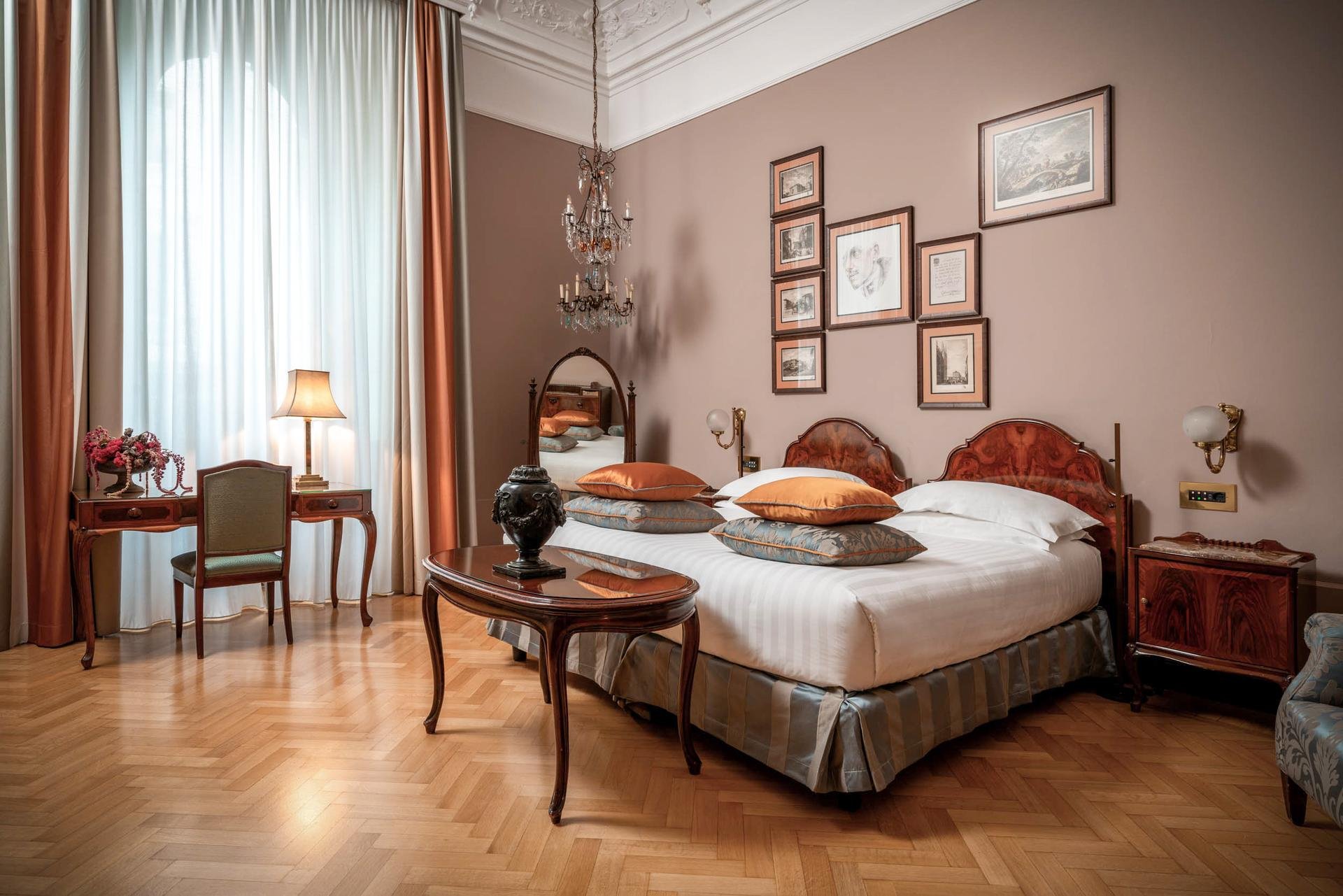 A vintage-style hotel room featuring a large bed with wooden headboard, multiple decorative pillows, a wooden side table with a lamp, a full-length mirror, and a gallery wall with framed artwork. The room has tall windows with sheer and curtain drapes, elegant crown molding, a chandelier, and a wooden parquet floor.