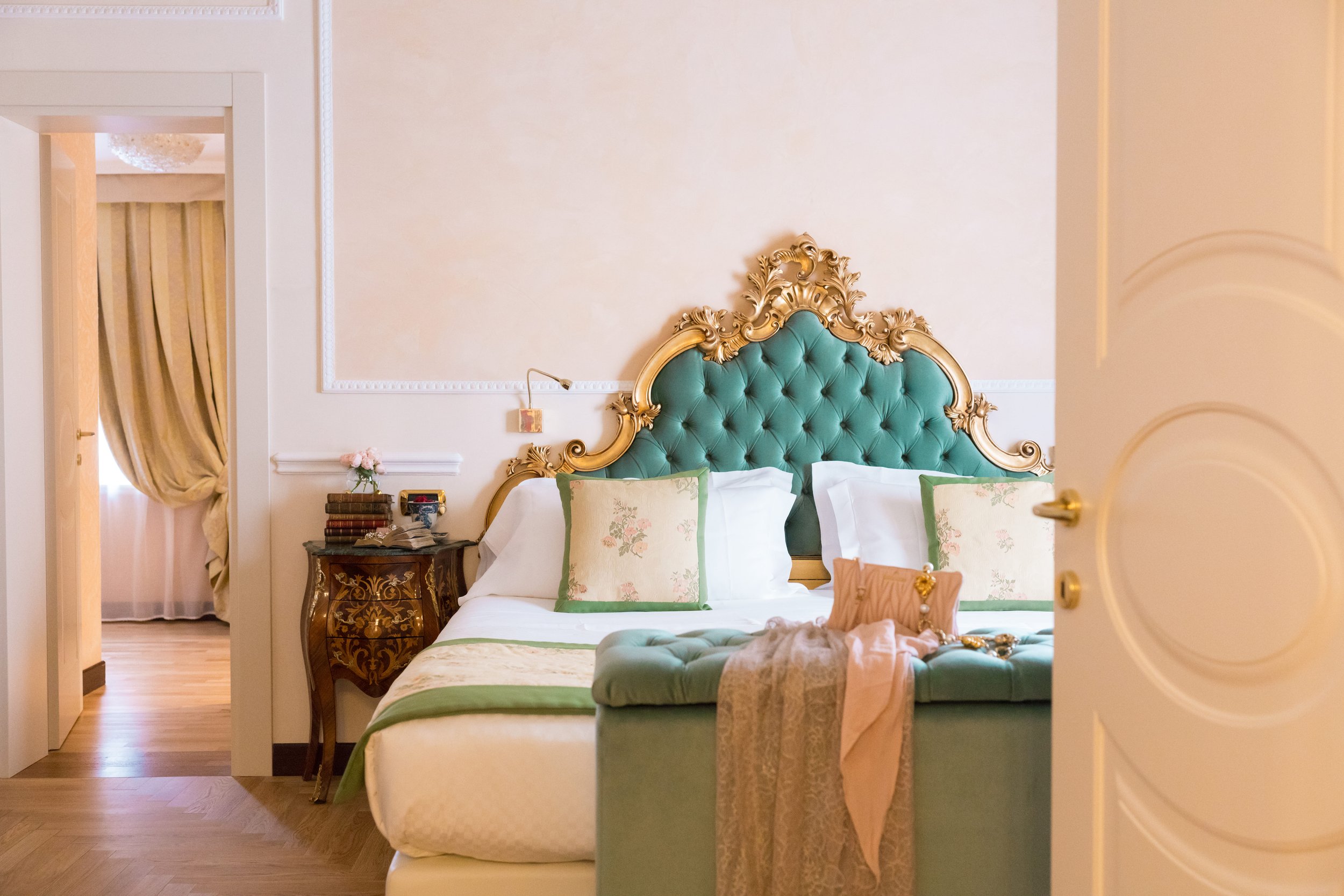 A luxurious bedroom with a green tufted headboard trimmed in gold, white bedding with green and floral pillows, a dark ornate nightstand with a small stack of books and a vase of pink flowers, and cream-colored curtains in the background. Part of a doorway with a gold doorknob is visible in the foreground.