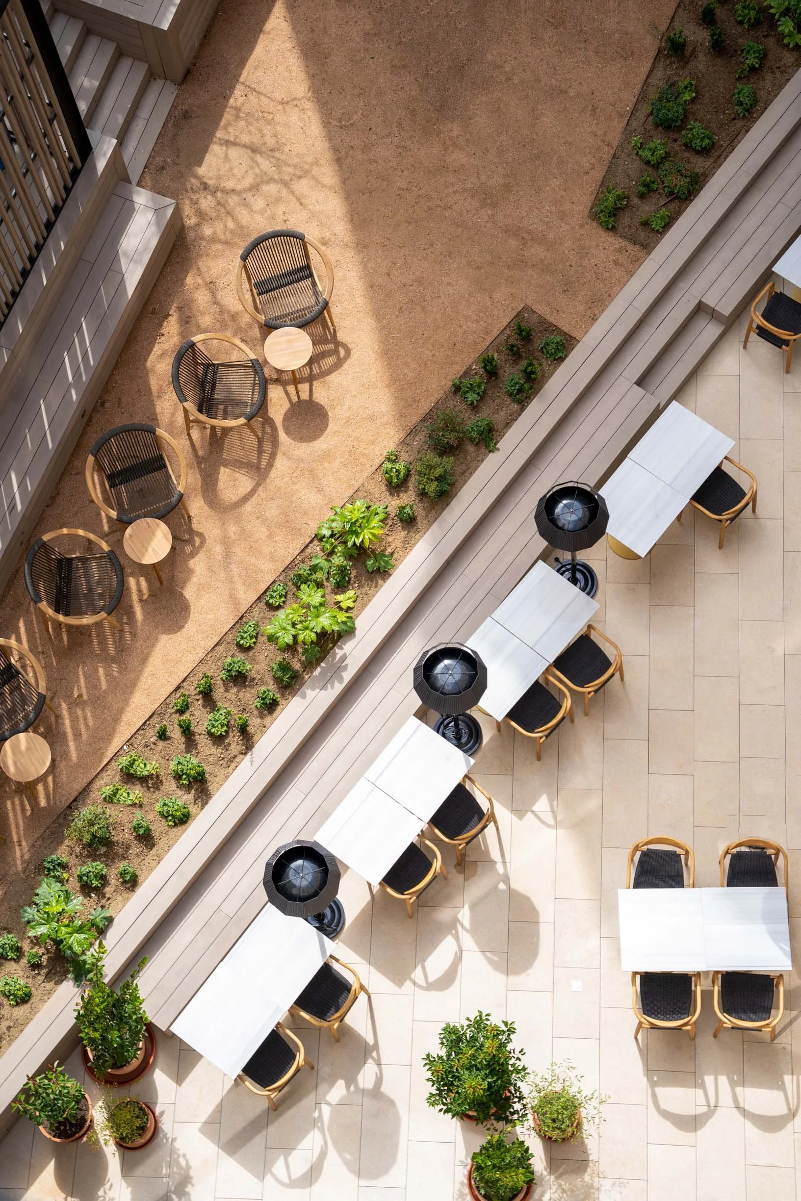 Top-down view of an outdoor patio area with white tables and black chairs, surrounded by potted plants and small garden beds, with a sandy ground area.