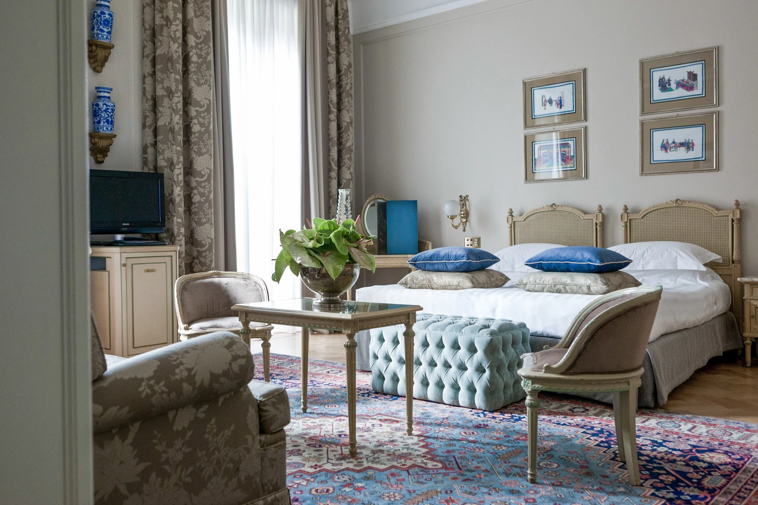 A bedroom with a double bed, adorned with blue and beige pillows, and a matching beige headboard. A large window with floral curtains allows natural light into the room. A bedside table with a lamp, a mirror, and decorative items is visible. There is