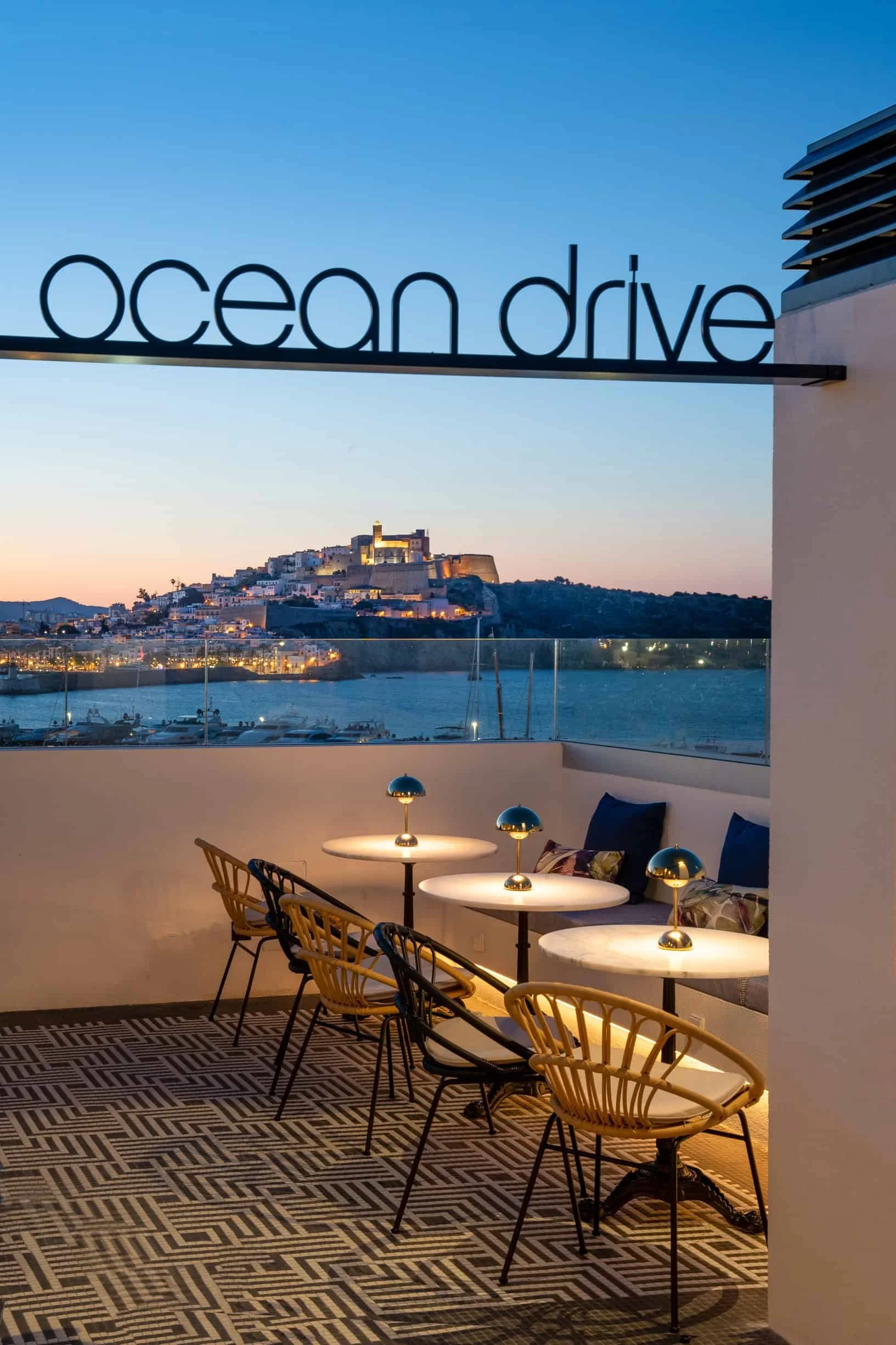 View of a restaurant outdoor seating area overlooking a coastal city with a historic fortress and harbor, under a clear evening sky.