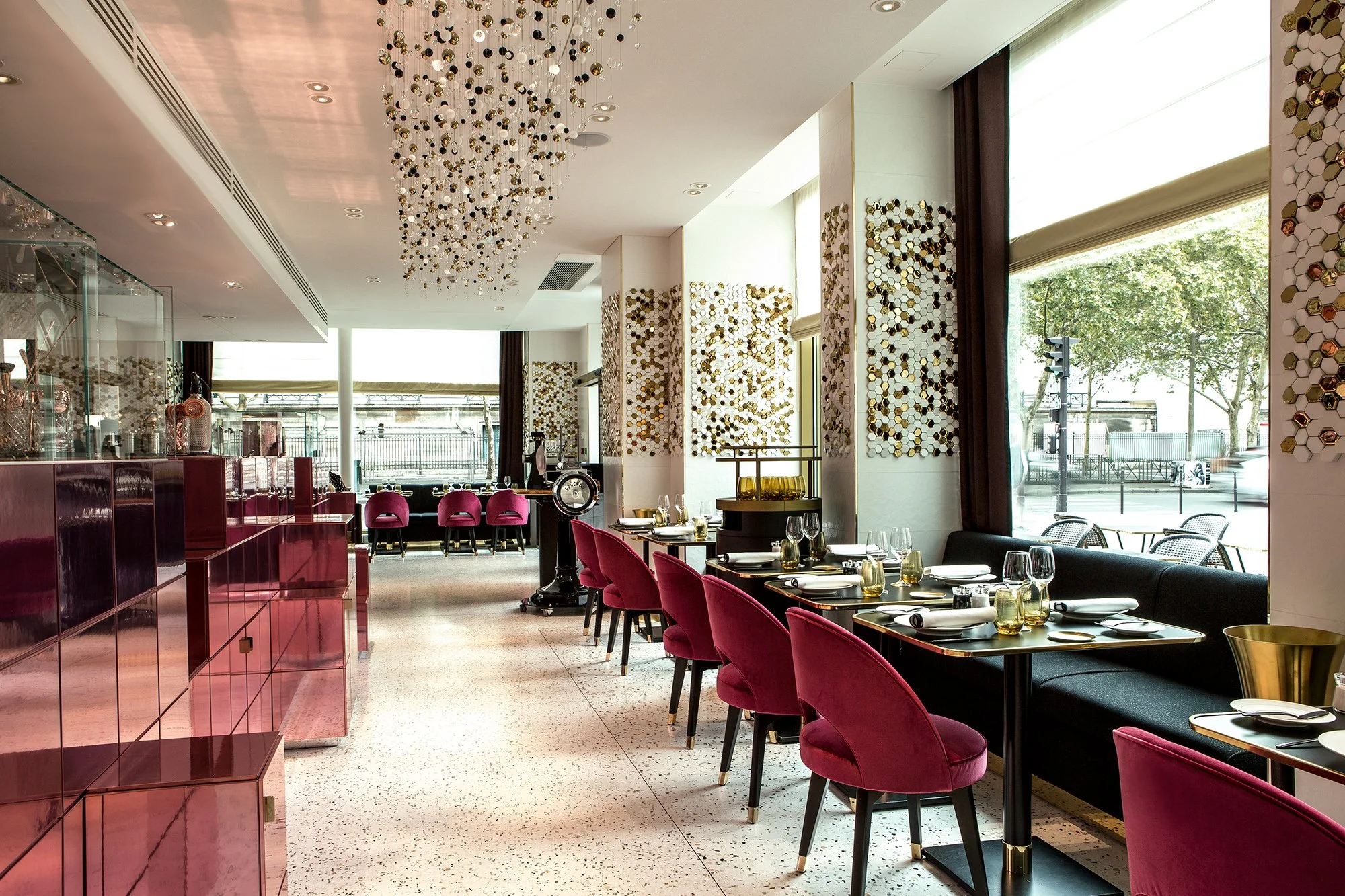 Upscale restaurant interior with pink velvet chairs, black tables, gold flatware, and large windows with trees outside. Modern decor with geometric wall art and a chandelier hanging from the ceiling.