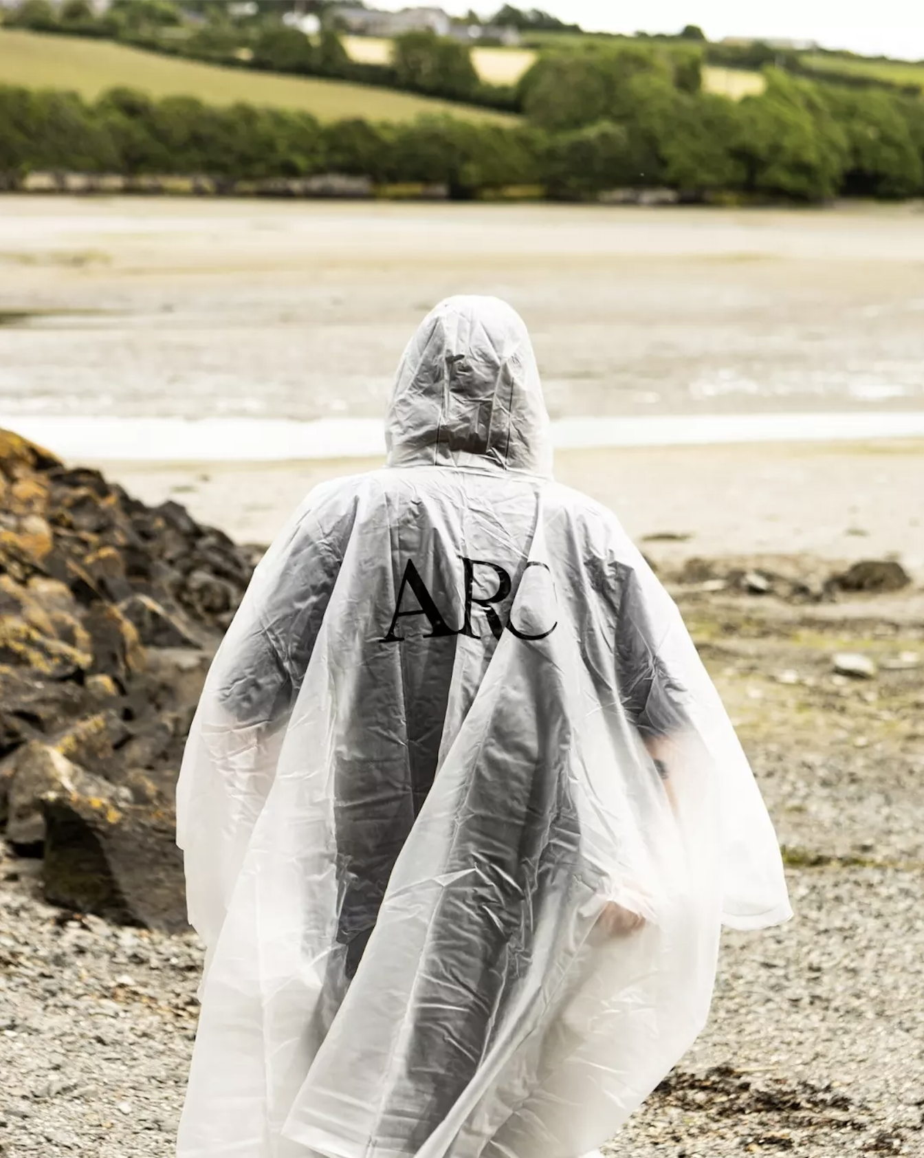 Person in a white protective suit walking on a rocky beach near water, with green trees and hills in the background.