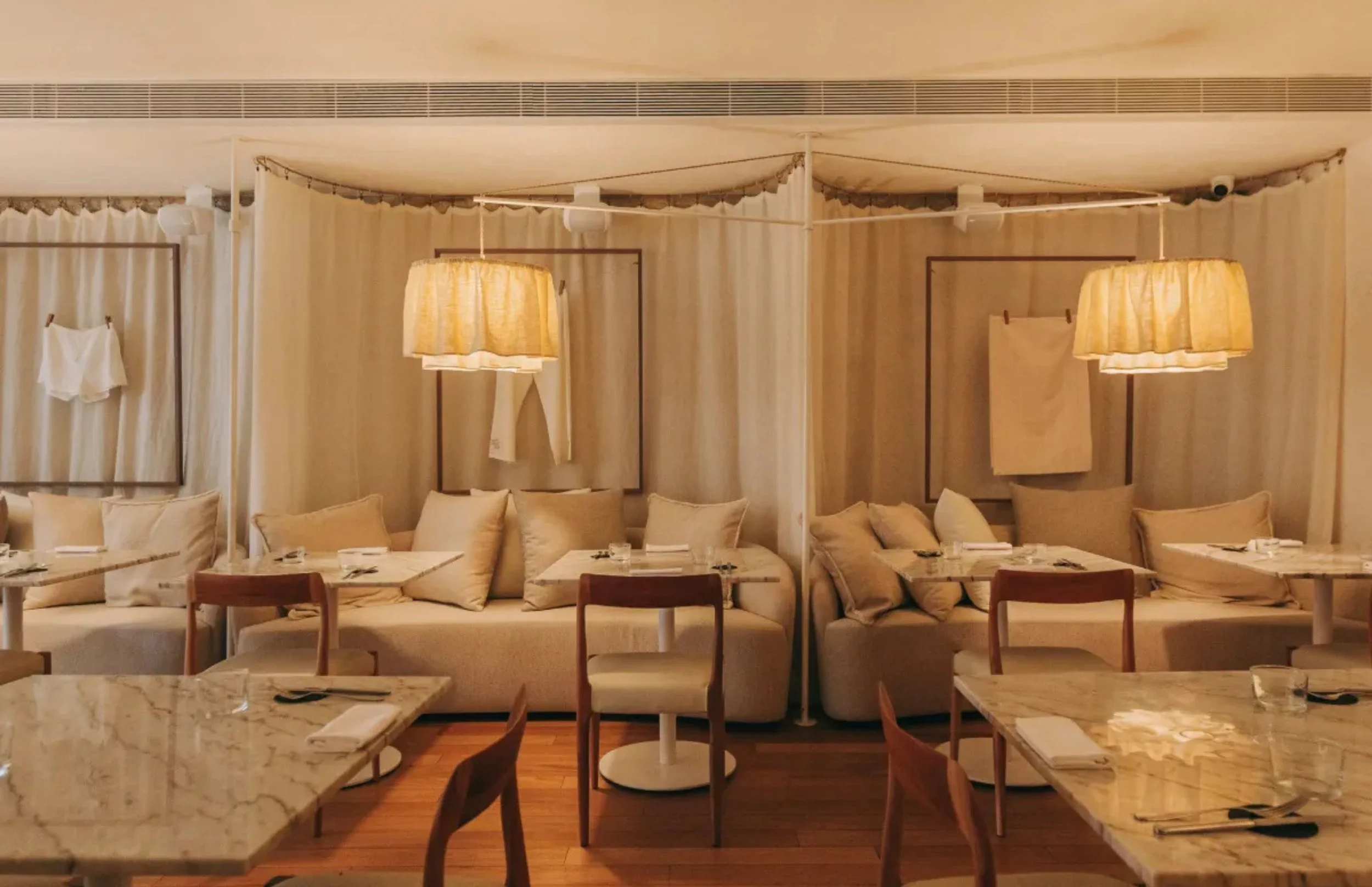 Interior view of a restaurant with marble tables, wooden chairs, beige cushions, hanging lamps, and curtains dividing the space.