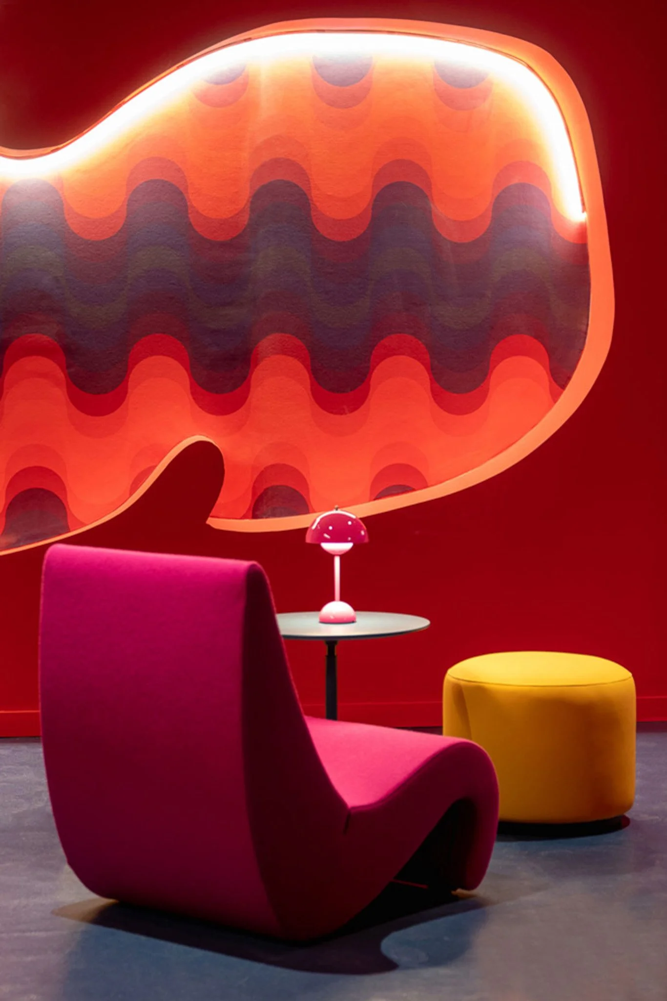Modern interior with a large red lounge chair, a yellow ottoman, and a pink table lamp, set against a vibrant red wall with abstract wall art.