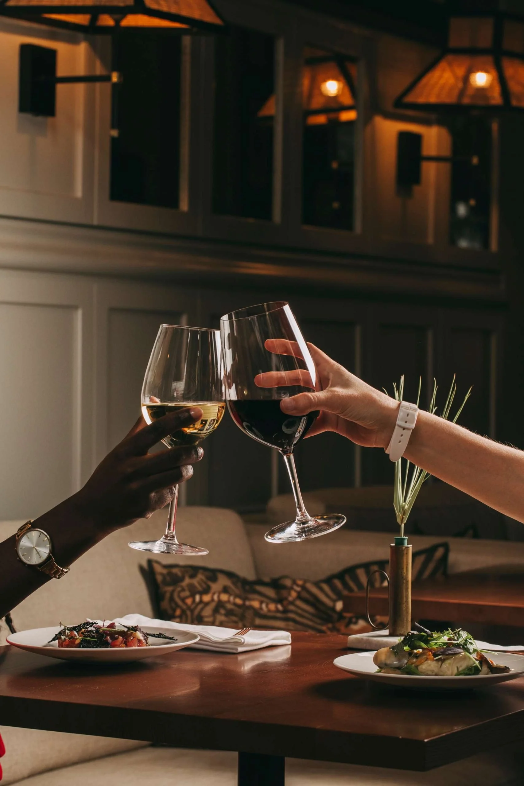 People clinking glasses of white and red wine in a cozy restaurant setting with plates of food on the table, warm lighting, and decorative elements in the background.