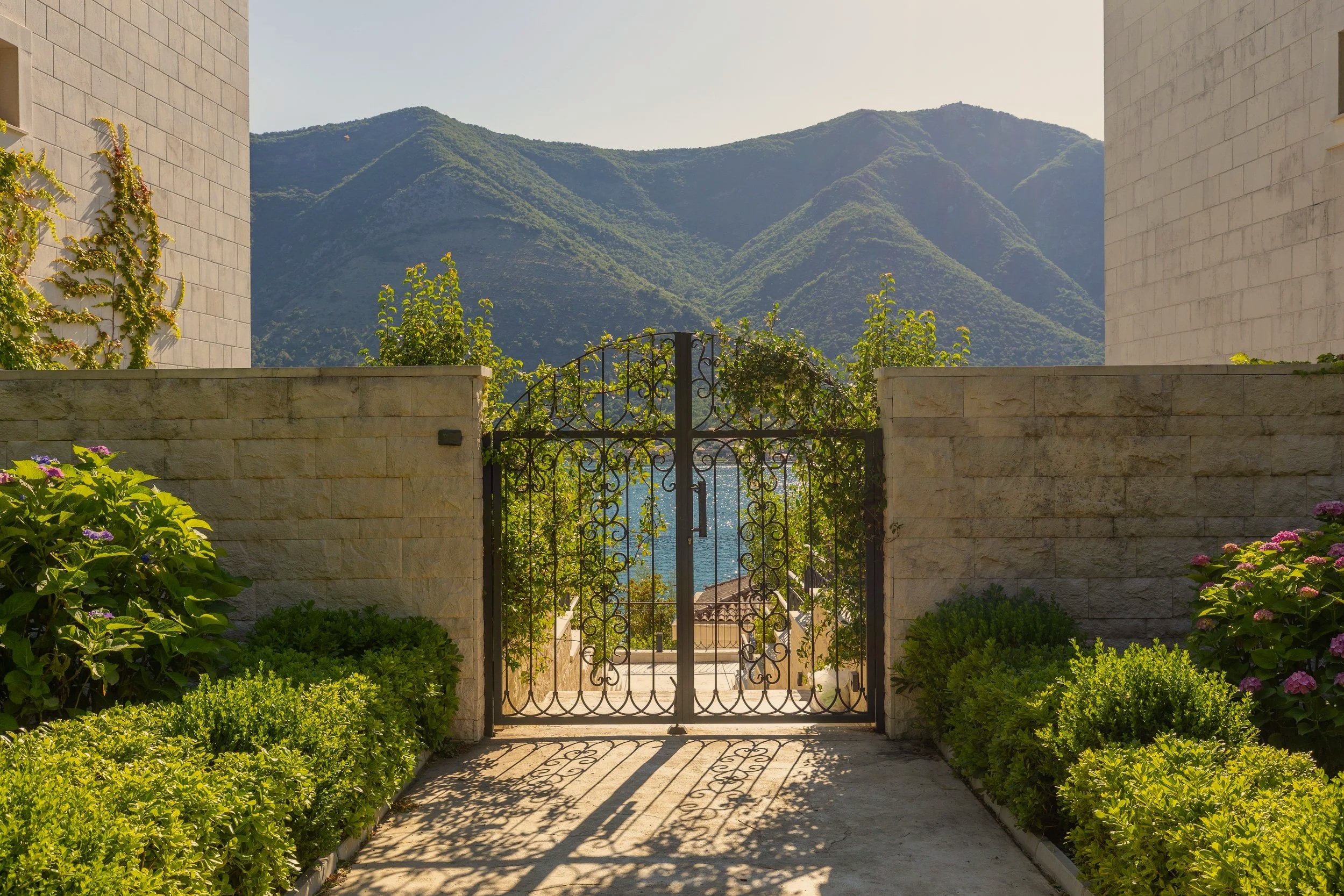 A view through a gate with a courtyard, surrounded by stone walls and lush greenery, overlooking green mountains in the distance.