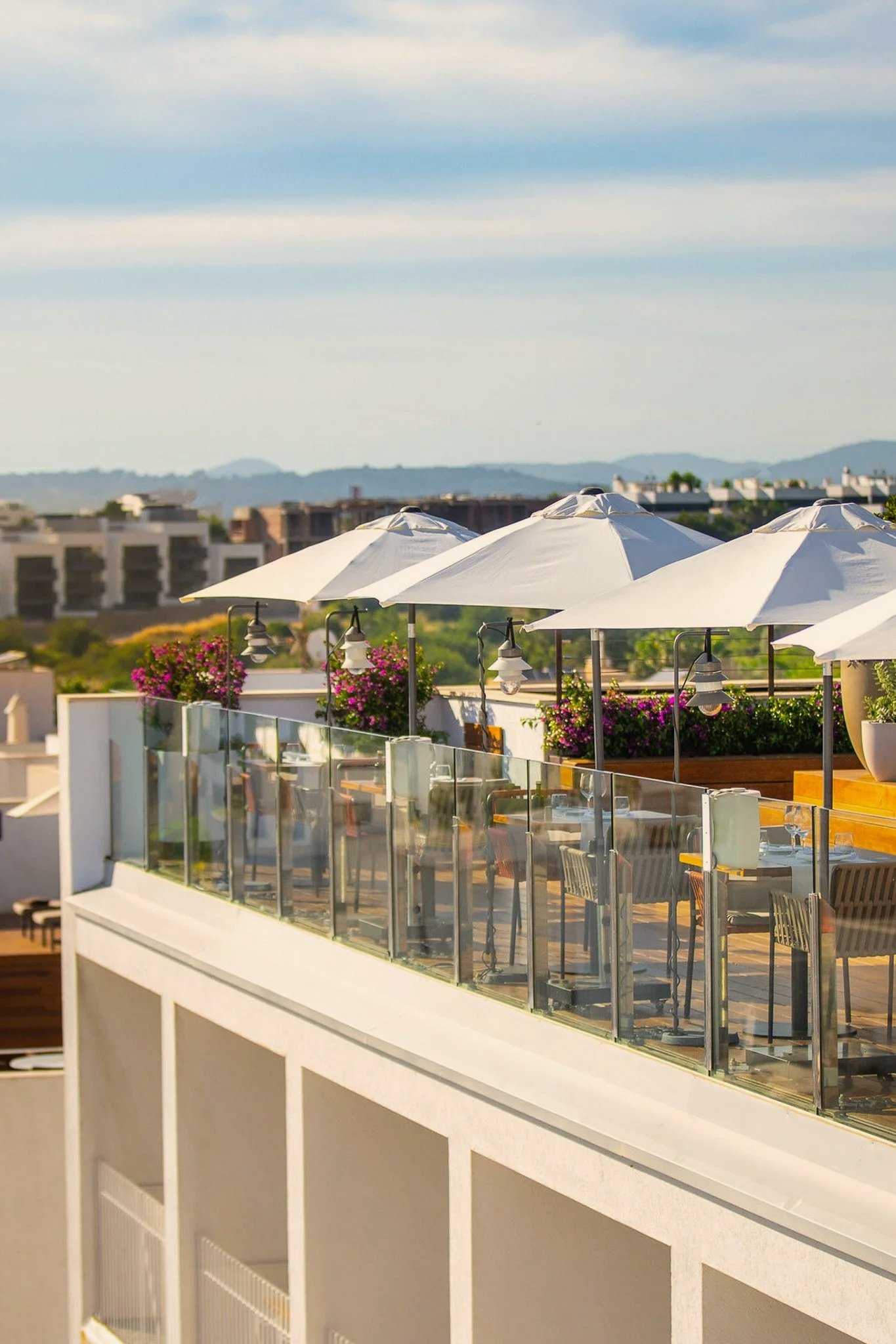 Outdoor terrace with white umbrellas, pink flowers, and glass railing overlooking a cityscape with buildings and hills in the distance.