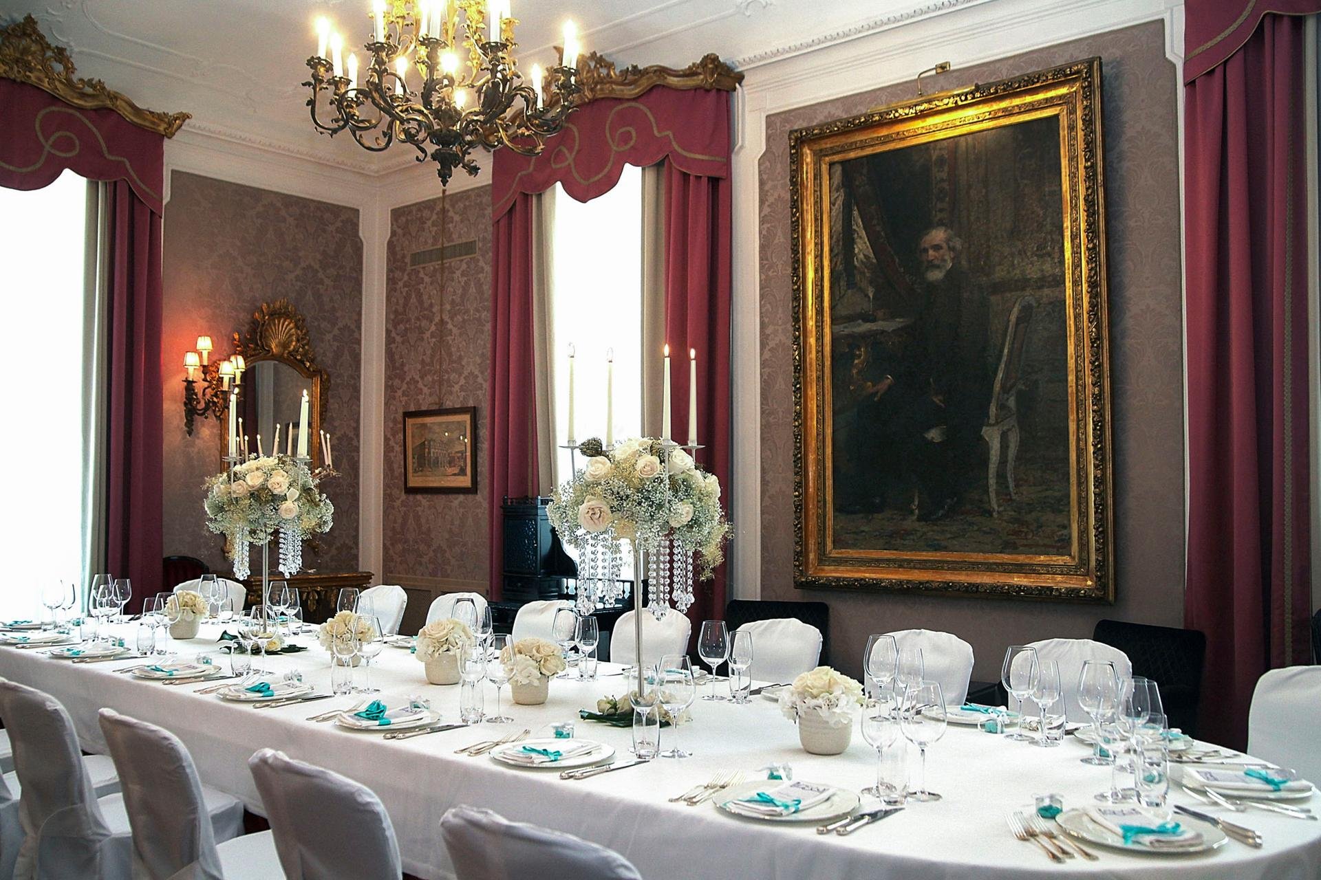Elegant banquet room with a long white table set for a formal event, decorated with white flowers and teal accents, surrounded by white chairs. The room features ornate pink curtains with gold trim, a large chandelier, candles, a decorative mirror, and a large framed portrait of a man.
