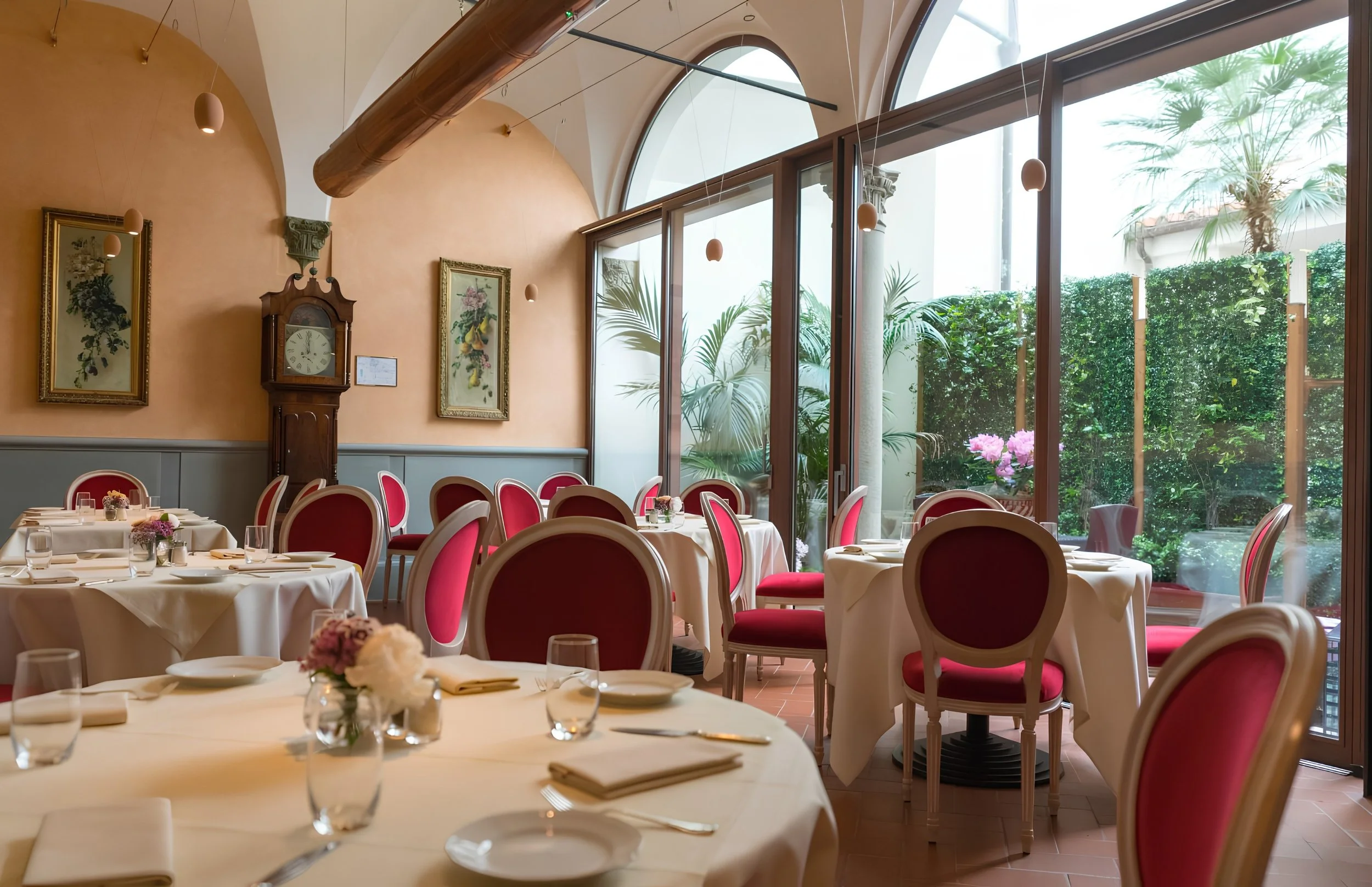 Elegant restaurant with large windows, potted plants, framed botanical artwork, antique clock, and set tables with white tablecloths, glassware, and flower centerpieces.